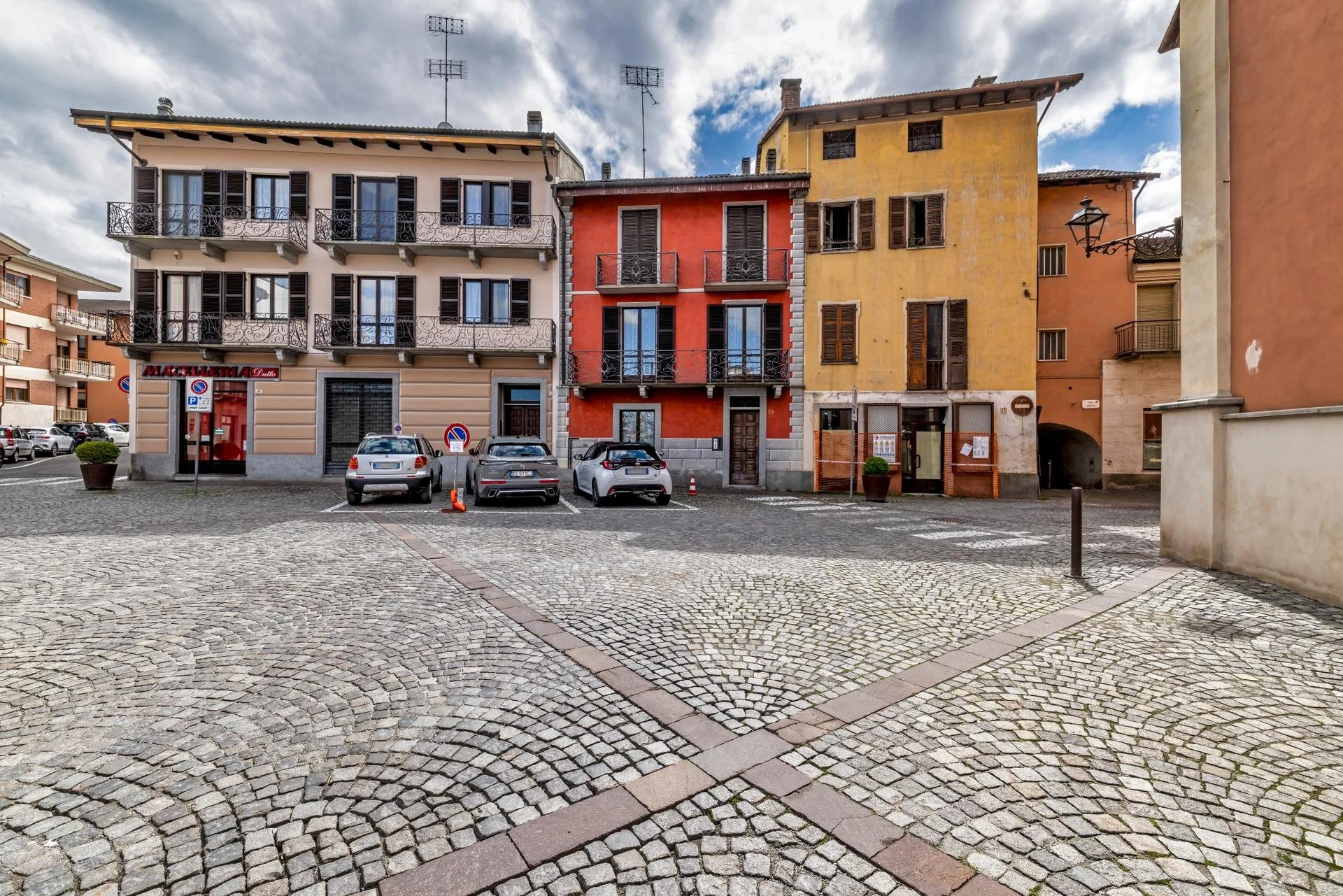European town square with cobblestone pavement, parked cars, and colorful multi-story buildings with balconies and shuttered windows under cloudy sky.