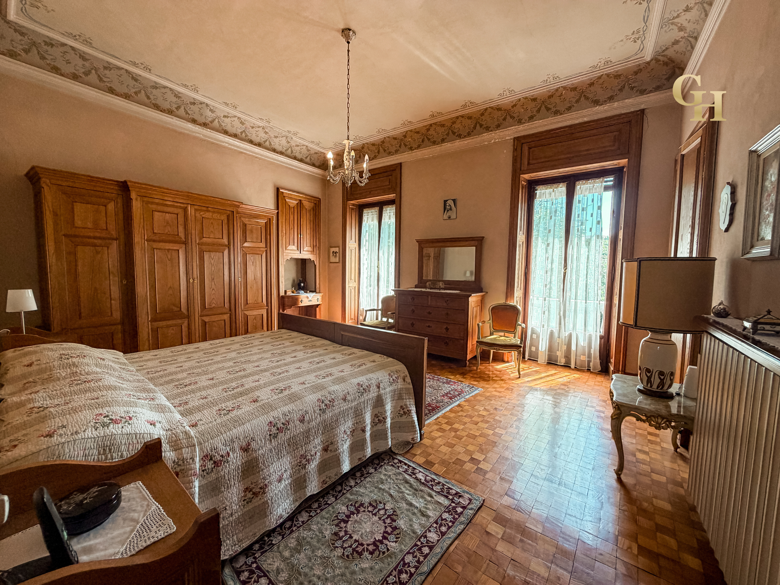 A vintage-style bedroom with wooden furniture, including two beds with floral bedspreads, a wardrobe, and a dresser. The room has large windows with sheer curtains, a chandelier hanging from the ceiling, and decorative items like framed pictures and 