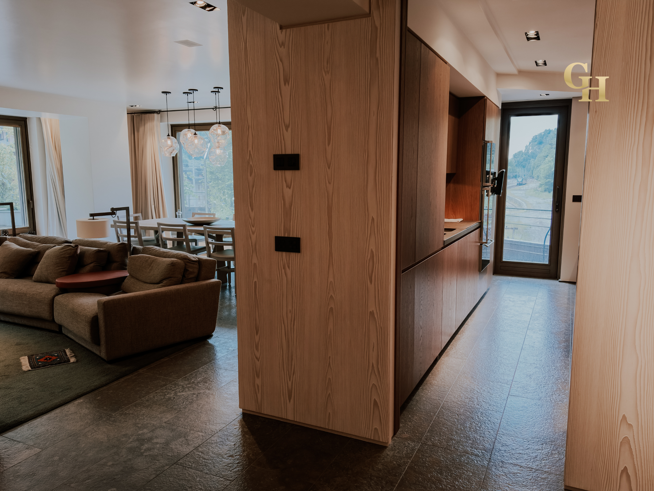 Interior of a modern living space with a brown sofa, dining table with chairs, and a partial view of a kitchen area with wooden cabinets, large windows showing an outdoor view, and contemporary ceiling light fixtures.