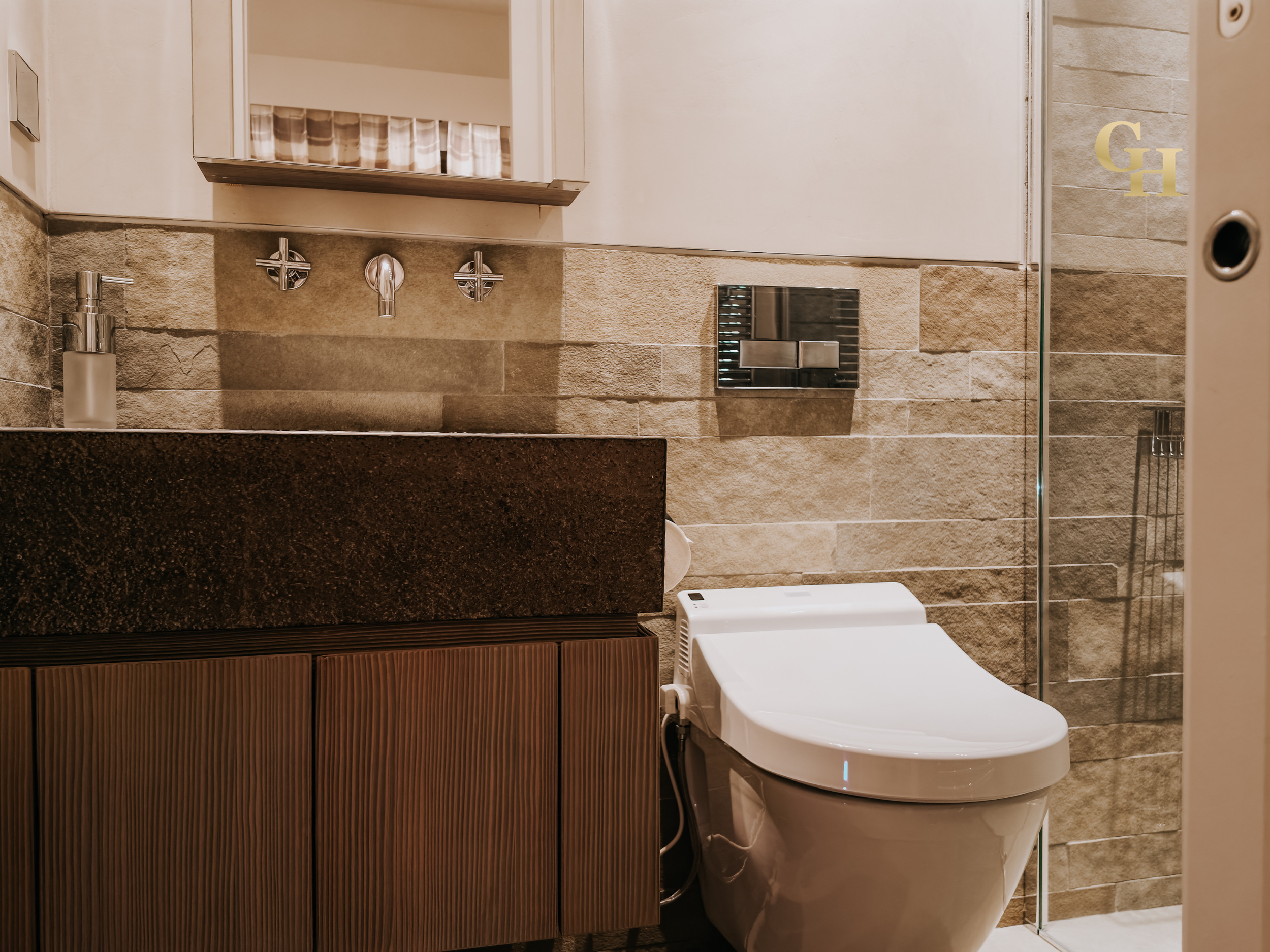 A modern bathroom with beige stone tile walls, a white toilet, a dark brown wooden vanity with vertical slats, a soap dispenser, a mirror cabinet, and a partial view of a shower with glass door.