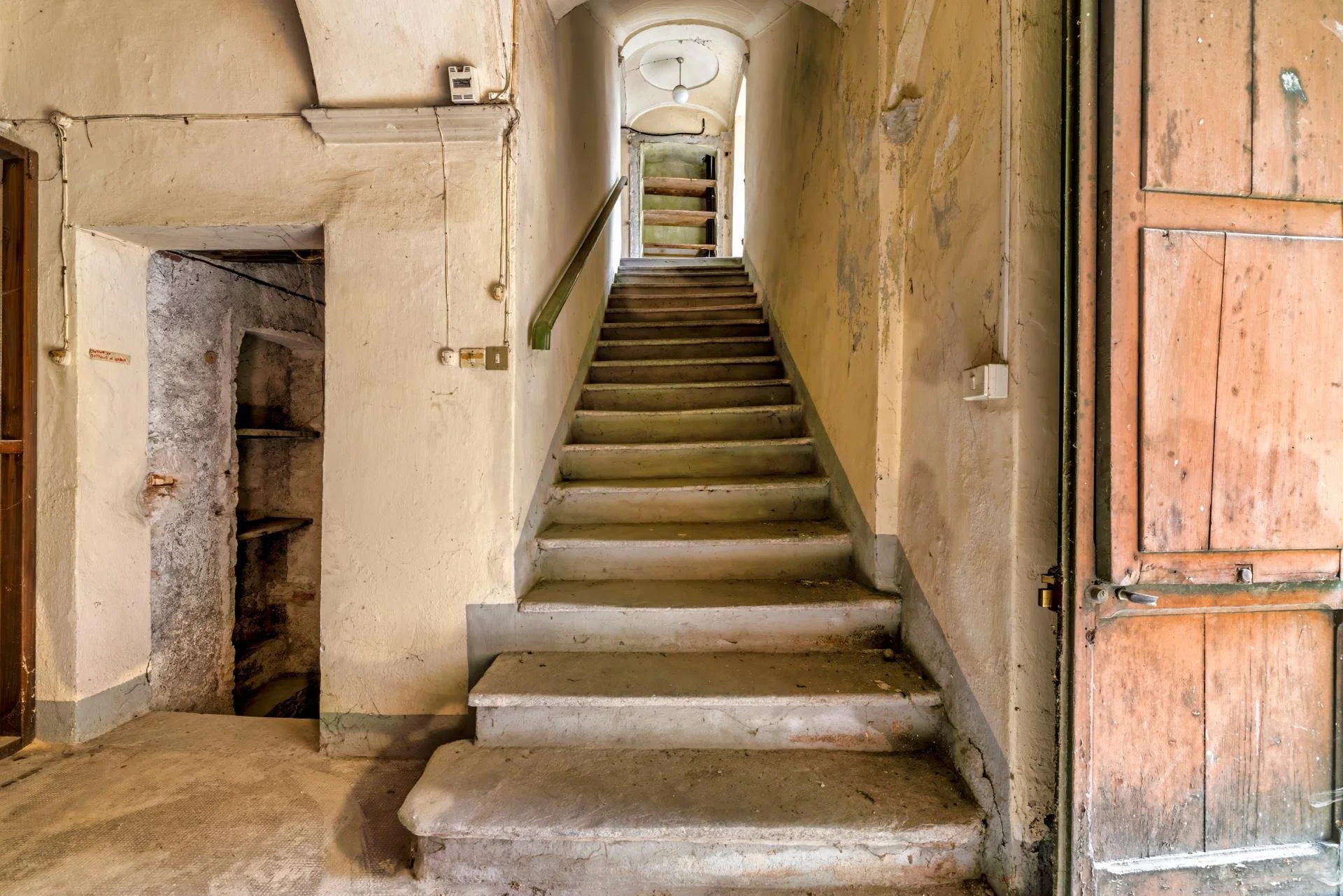 An old, worn staircase leading to an upper level in a dilapidated building with peeling paint and exposed wiring.