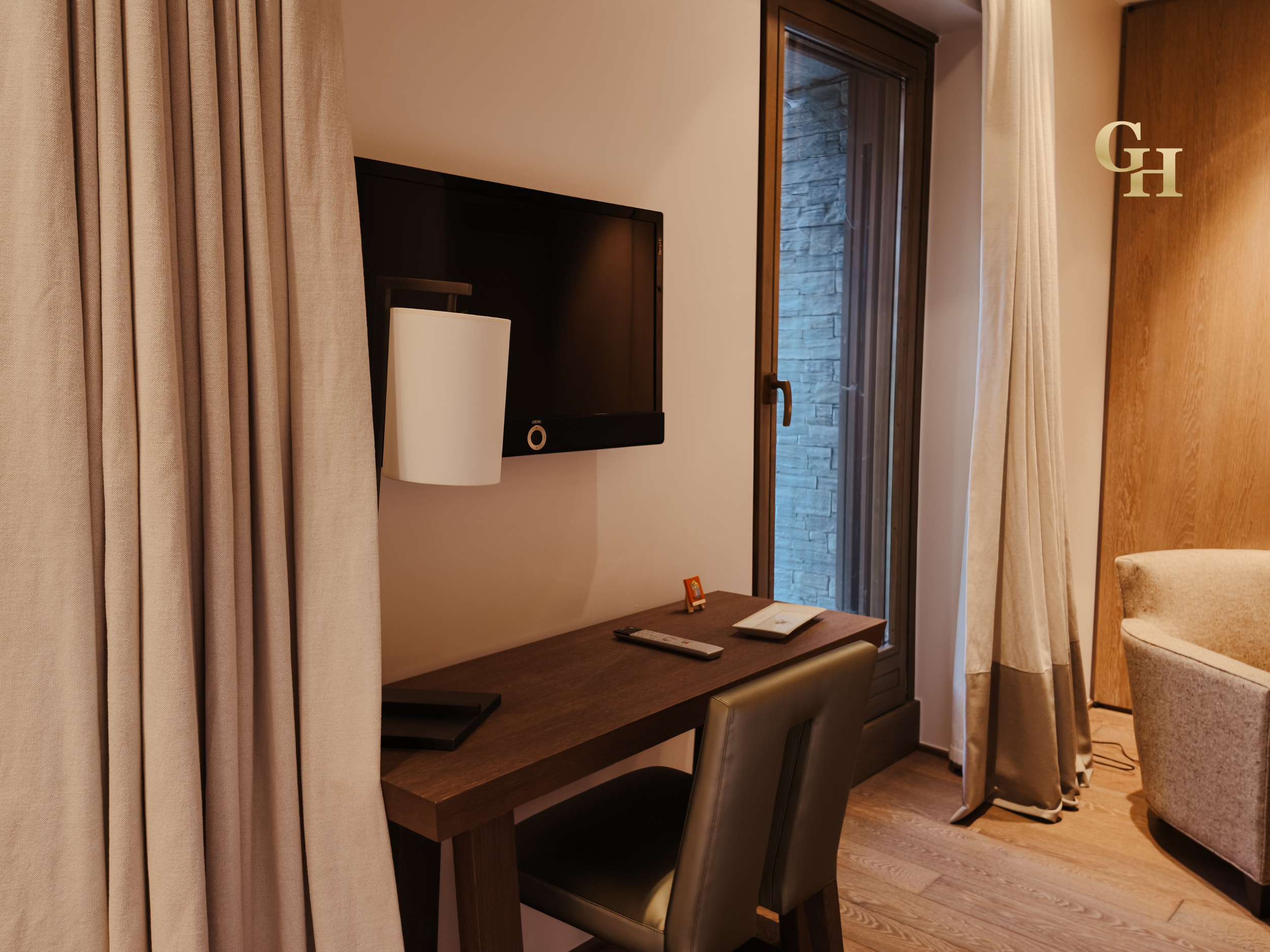 Interior view of a hotel room featuring beige curtains, a flat-screen TV, a desk with a remote control, a chair, a window with a stone wall outside, and part of a beige armchair.