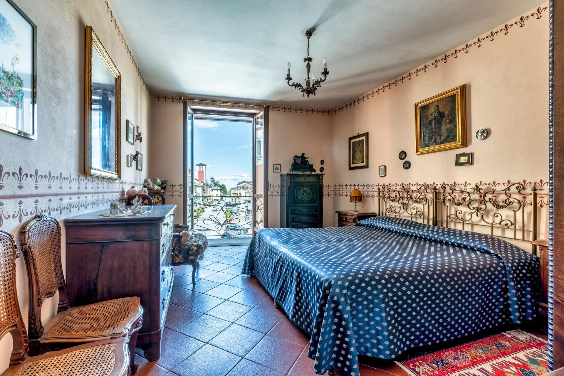 A vintage bedroom with a window showing a cityscape and blue sky. The room has a wrought iron bed with a blue patterned bedspread, antique wooden furniture, and framed artwork on the walls.