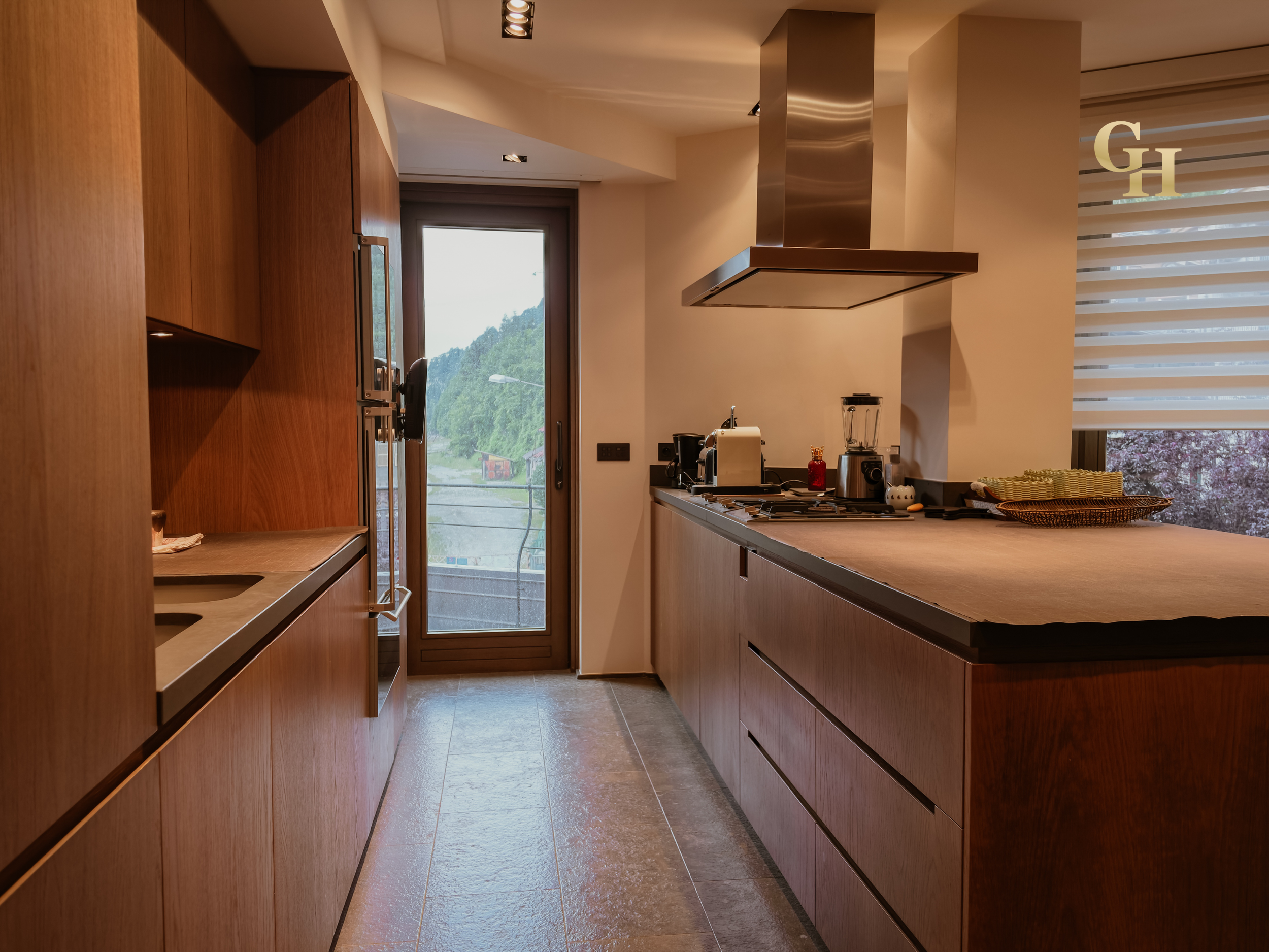 A modern kitchen with wooden cabinets, a countertop with appliances, and a large window showing a view of a green hill and cloudy sky.