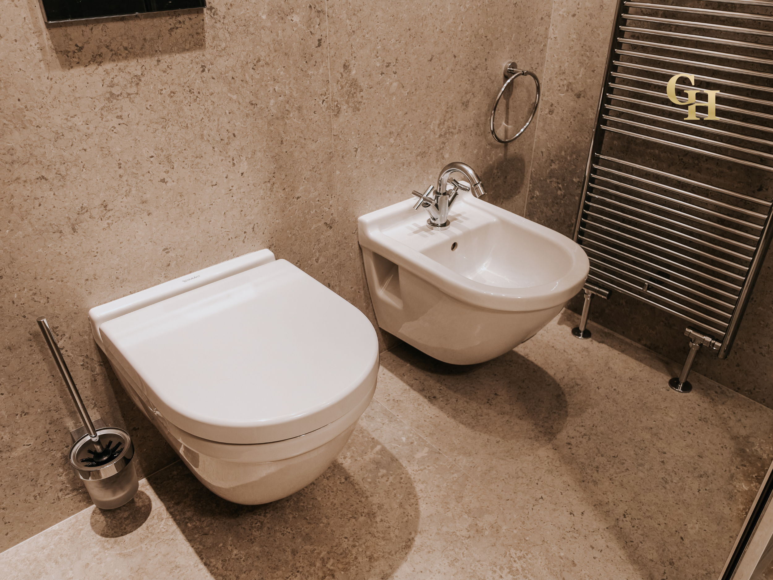 Bathroom with beige tiled walls and floor, featuring a white bidet with a silver faucet, a silver towel ring on the wall, and a modern white toilet with a silver toilet brush holder nearby. There is also a large metallic heated towel rack with a logo