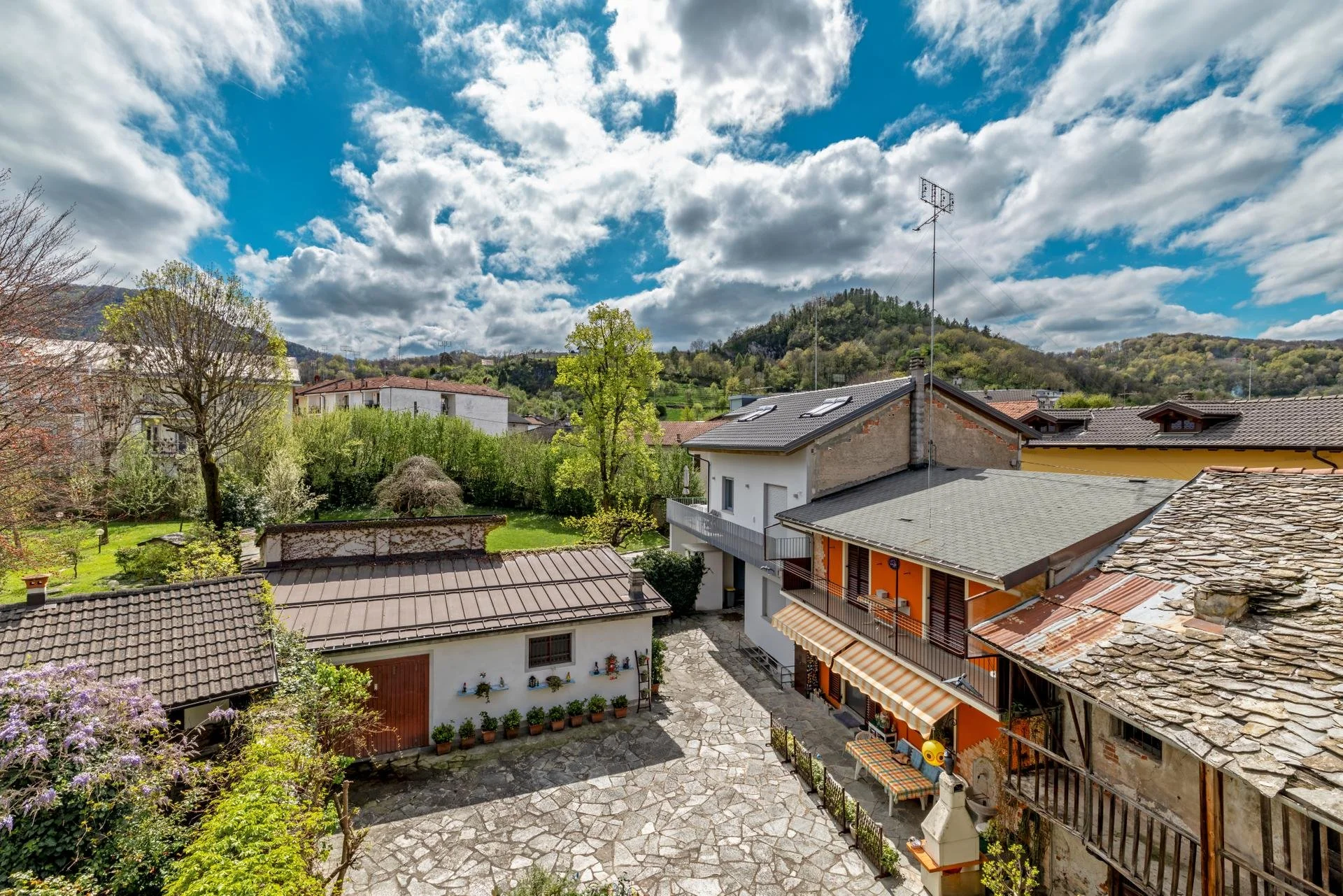 View of a residential backyard with paved patio, surrounding plants, and neighboring houses with hilly landscape and partly cloudy sky in the background.