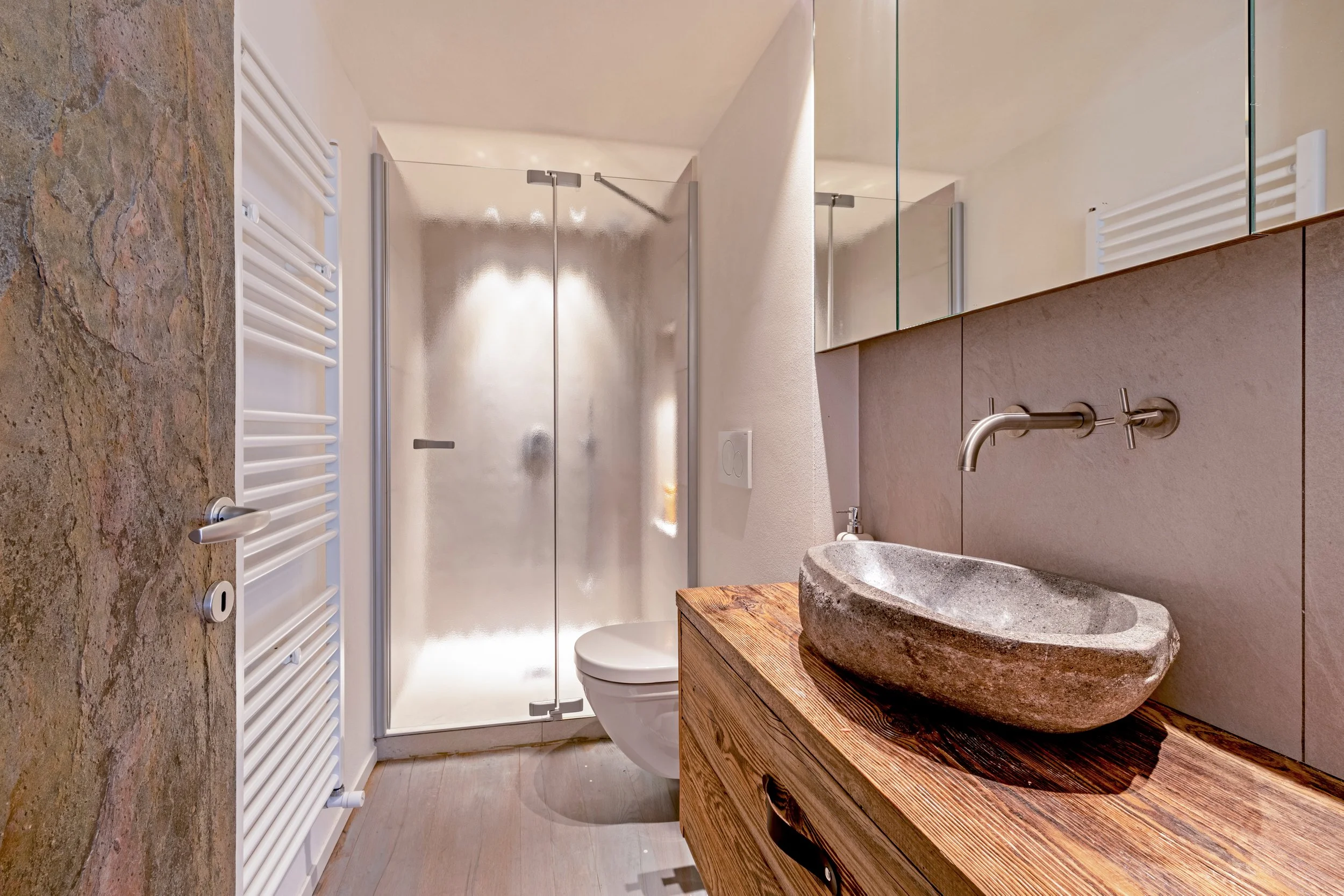 A modern bathroom featuring a stone vessel sink on a wooden vanity, a frosted glass shower enclosure, a white toilet, and a heated towel rack on the wall.
