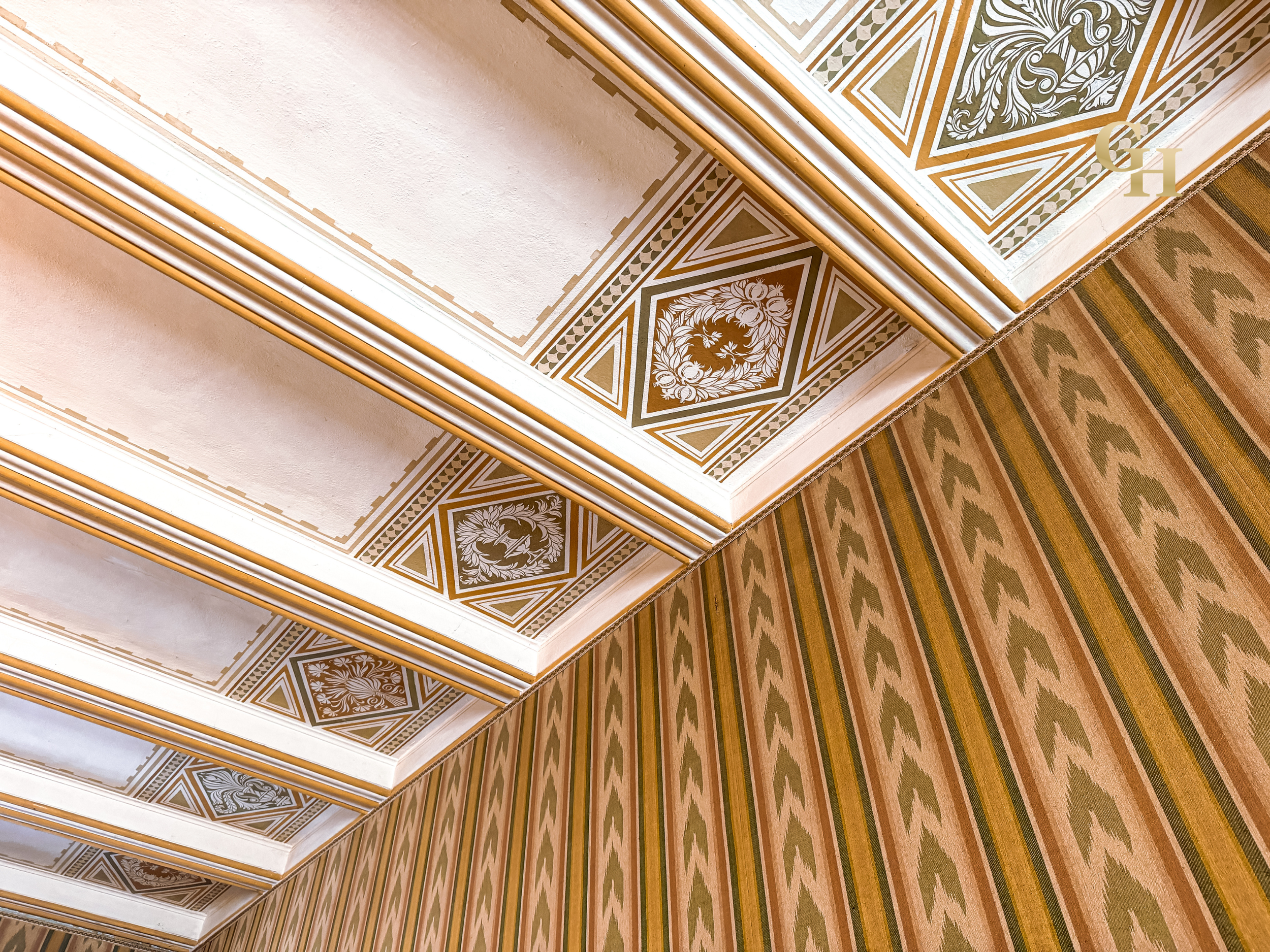 Detailed view of ornate ceiling moldings and decorative wall patterns in a historic building, featuring gold, green, and beige colors with intricate floral and geometric designs, and a patterned carpet on the floor.