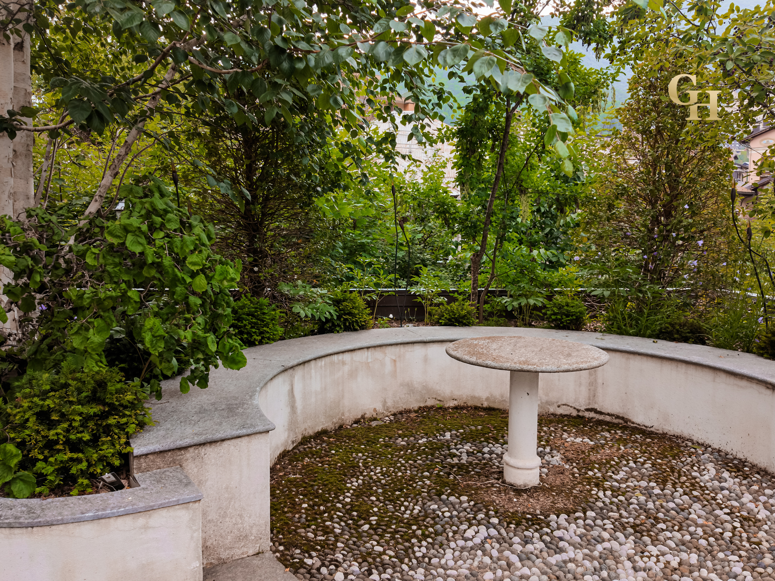 A curved concrete bench with a round concrete table in the center, surrounded by green shrubs and trees in a garden, with a pebble ground and a white wall on the left side.
