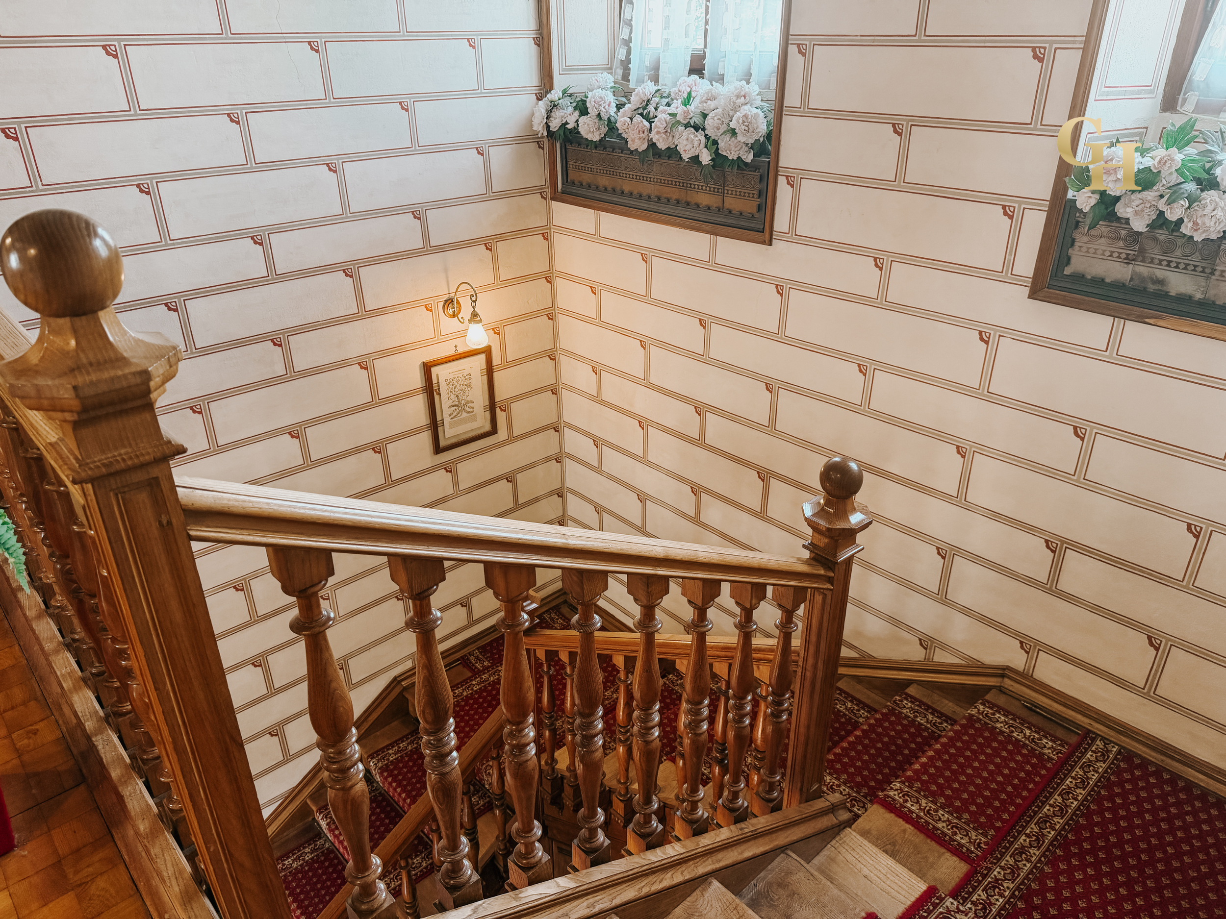 Wooden staircase with carved balusters and a patterned red carpet, leading down in a house with cream-colored walls decorated with framed pictures and floral arrangements near windows.