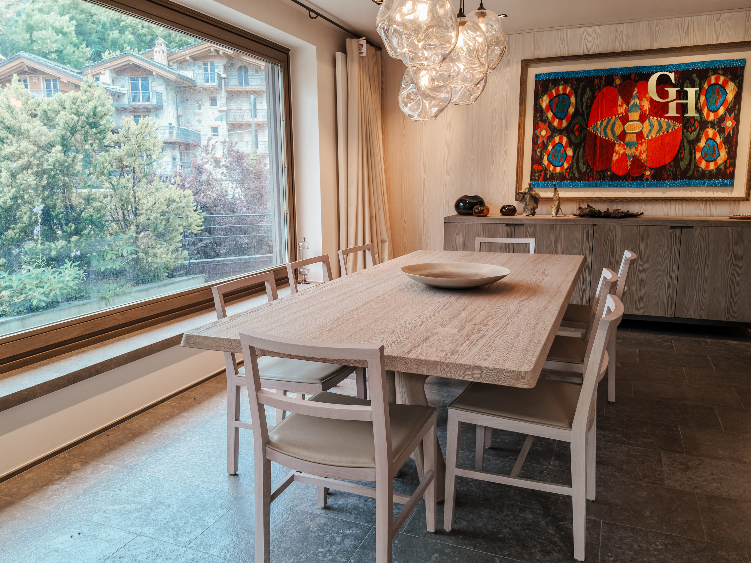 Modern dining room with large window showing trees and neighboring buildings, light wood table with a wooden bowl, white chairs, artistic glass chandelier, and colorful abstract painting on the wall.