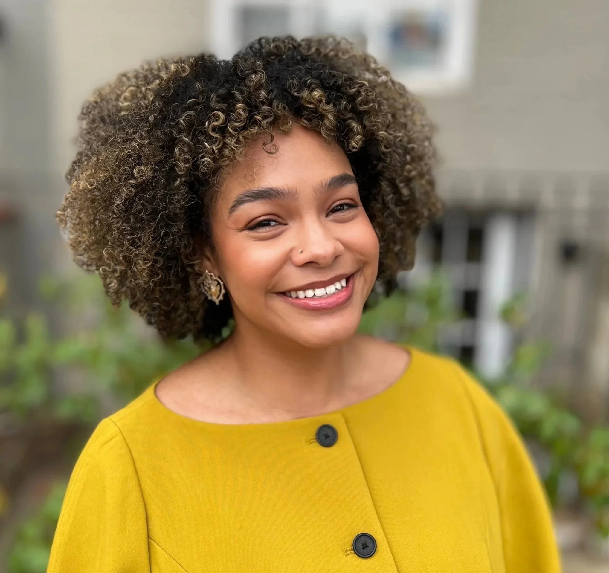 Close-up of a smiling woman with curly hair, wearing a yellow top with black buttons, standing outdoors with blurred greenery and a fence in the background.