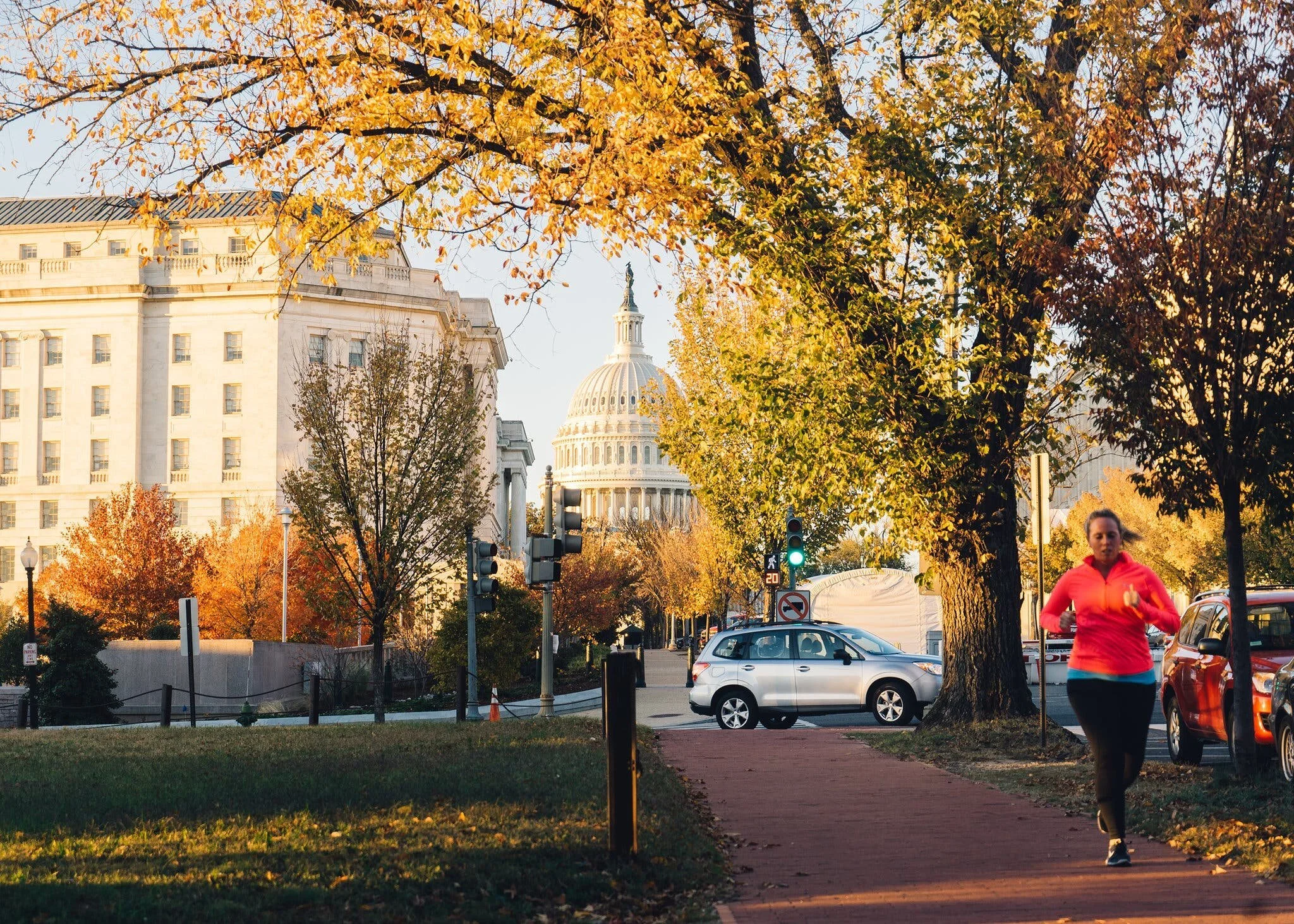 View of the U.S. Capitol building seen through trees with yellow and orange fall leaves. A woman in a pink jacket is jogging on the sidewalk in the foreground. Several parked cars are along the street, and traffic lights are visible.