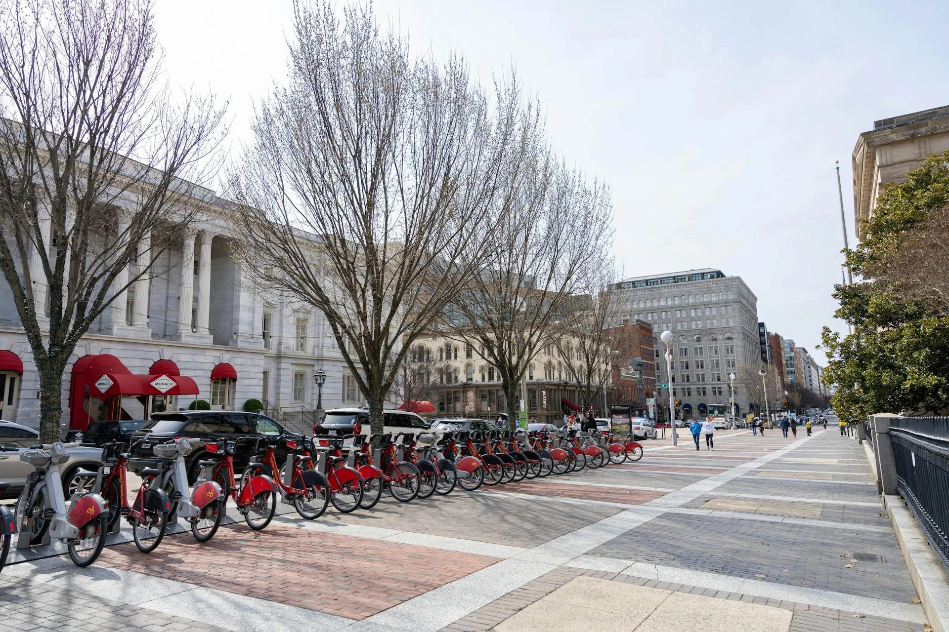 City street scene with a row of rental bikes in the foreground, leafless trees, historic buildings, and people walking on the sidewalk on a clear day.
