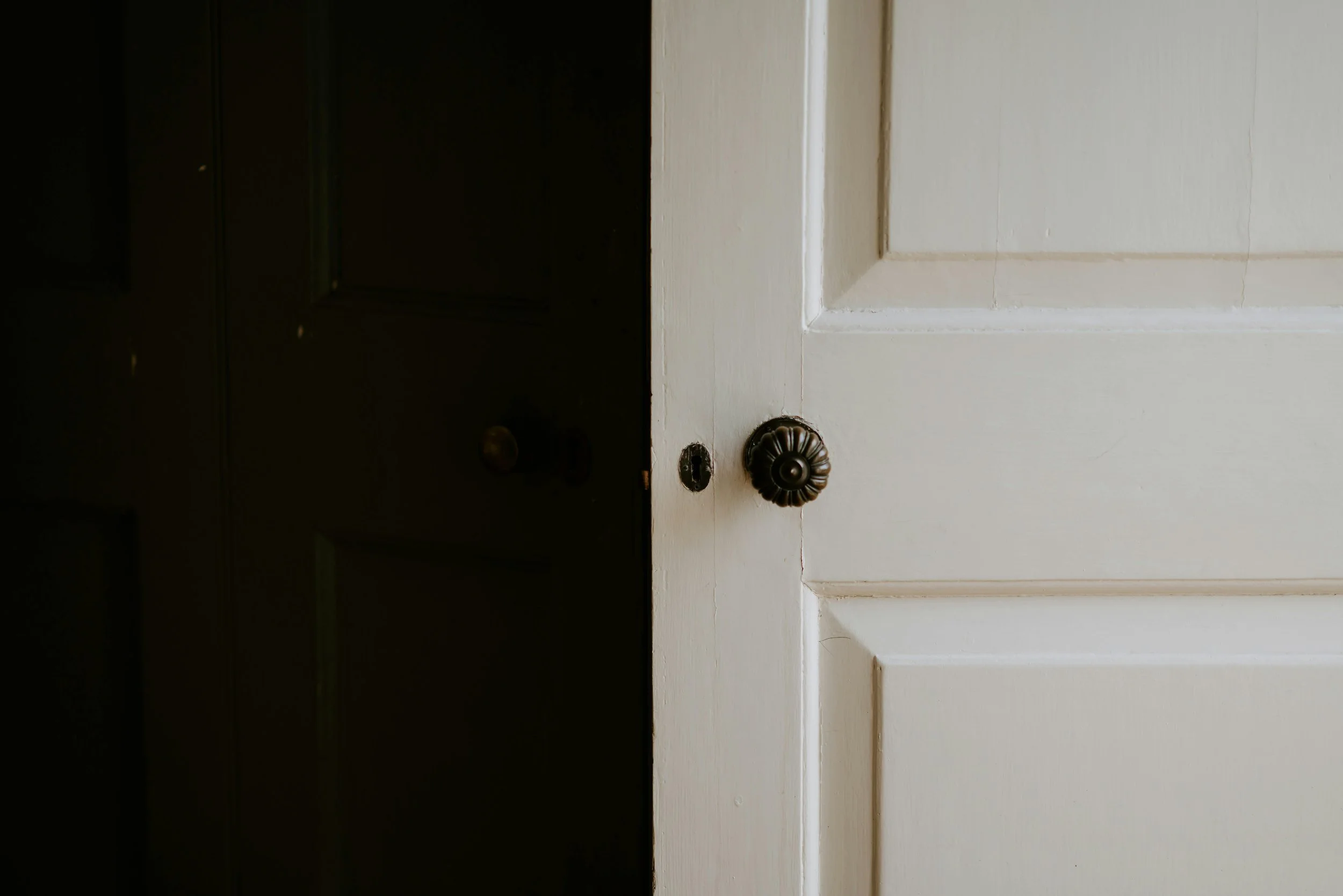 Close-up of a partially open white paneled door with an ornate dark doorknob, revealing a dark room behind it.