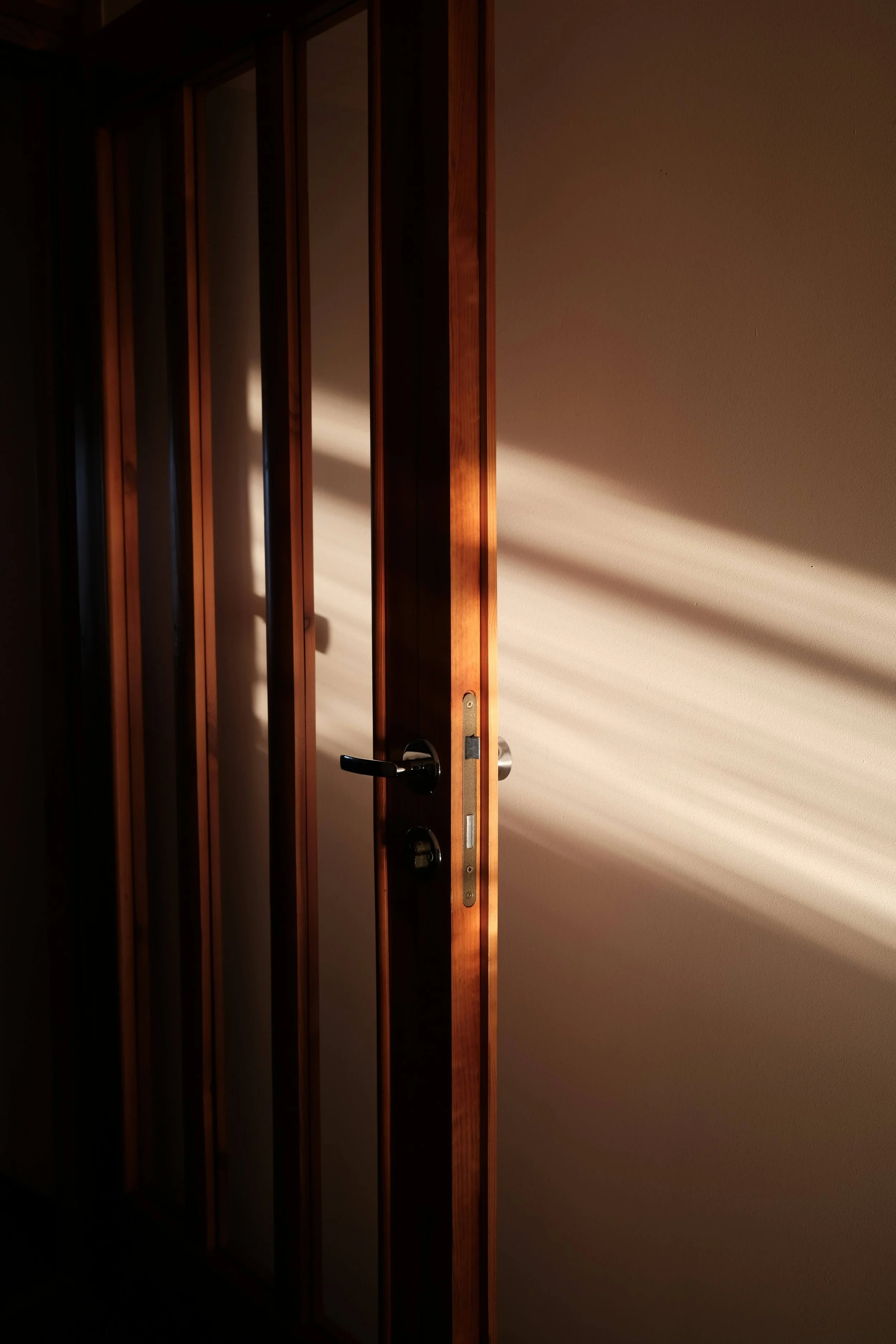 Partially open wooden door with black handle and deadbolt casting shadows on a sunlit beige wall