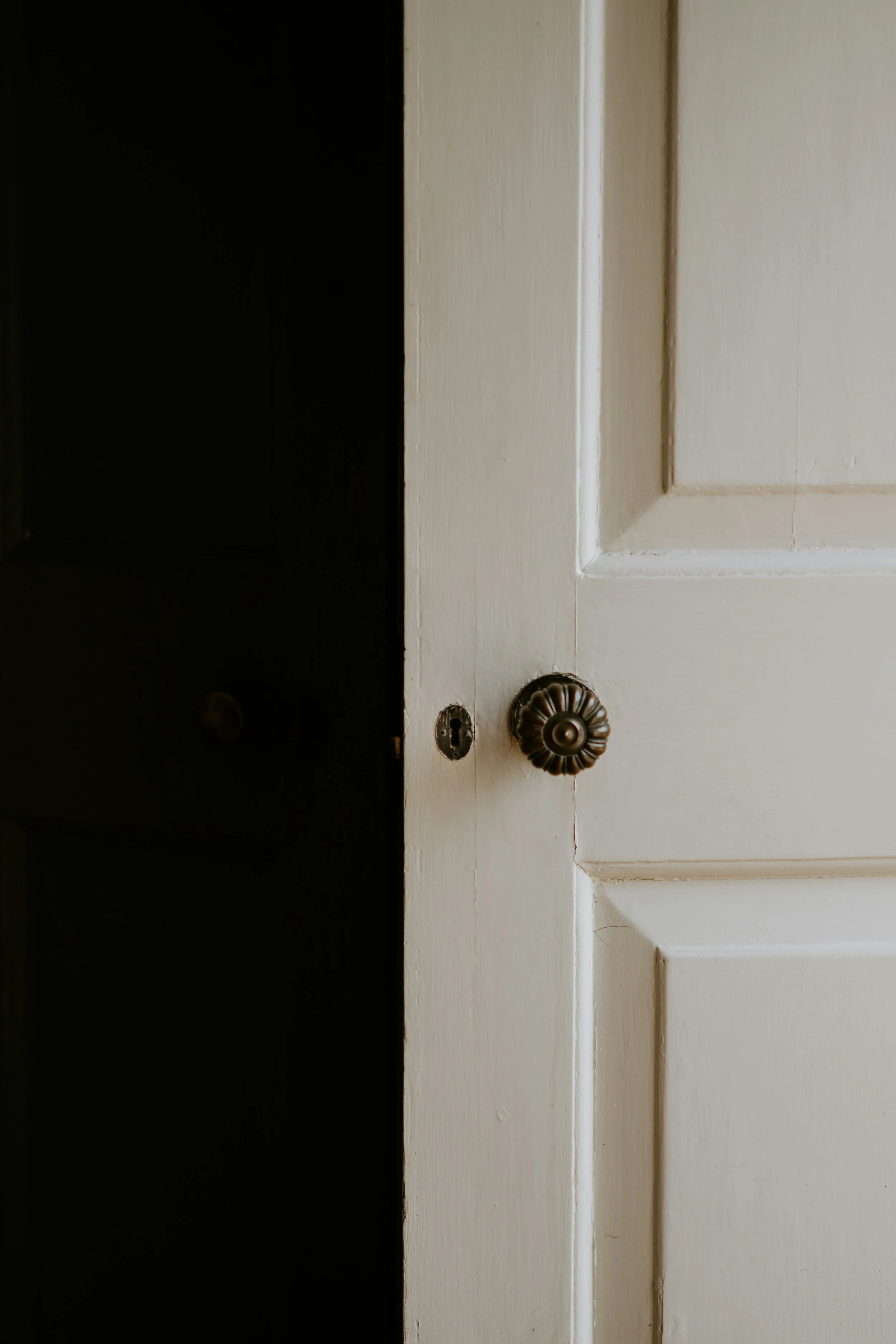 Close-up of an open white paneled door with an ornate brass doorknob and keyhole leading into a dark room