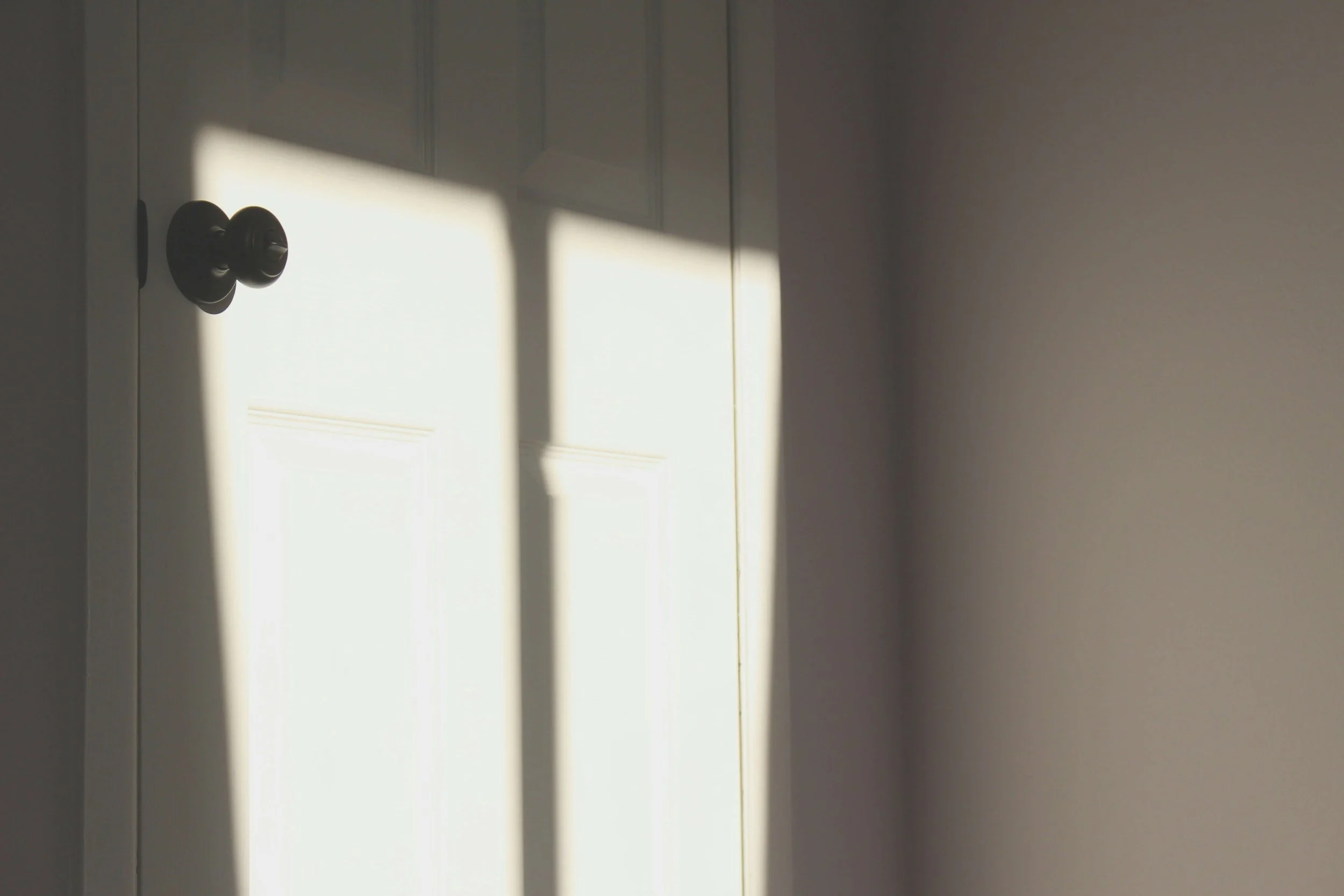 Sunlight casts windowpane shadows across a white paneled door with a dark round doorknob.