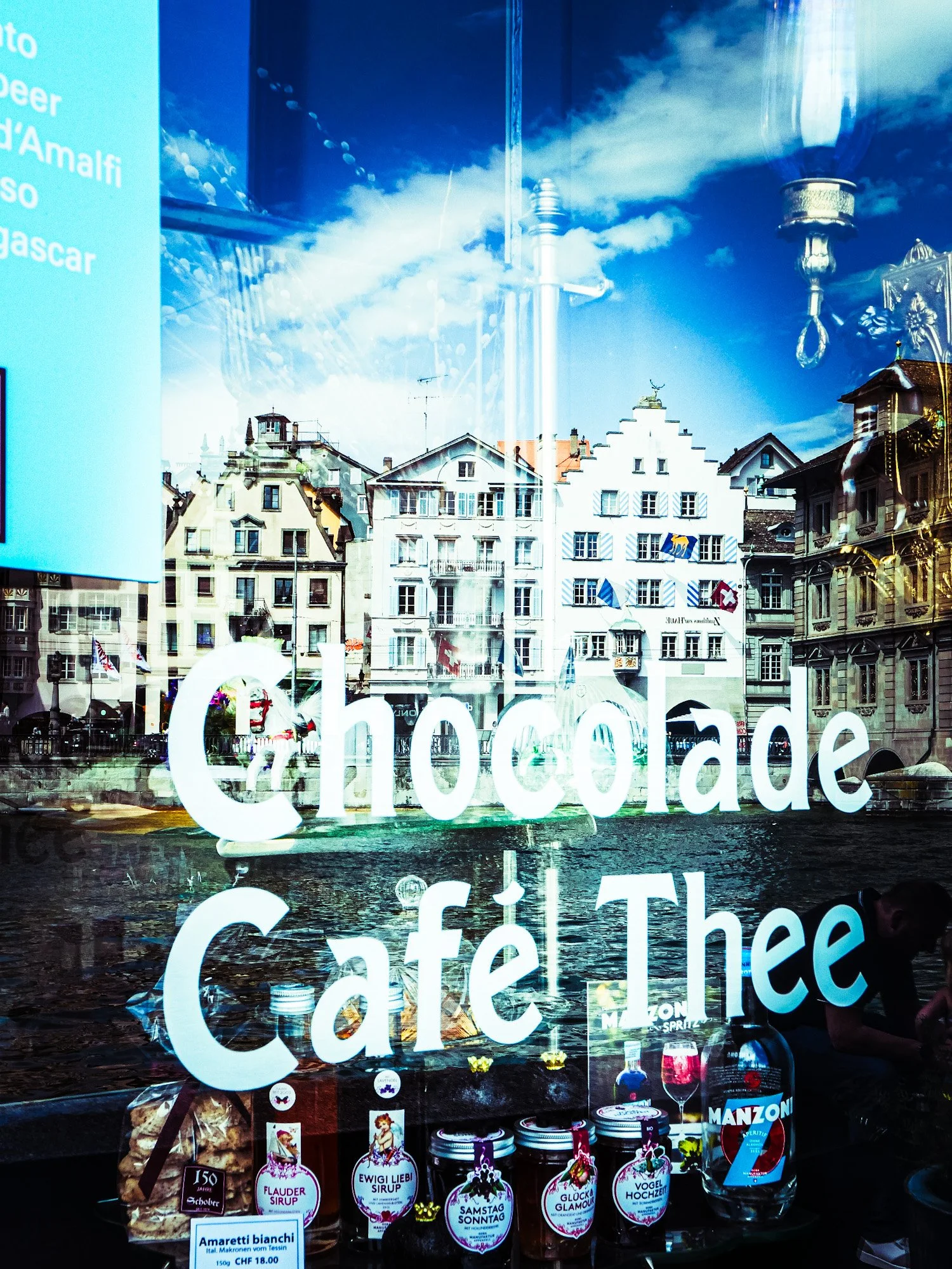 Eine Schaufensteransicht mit Gebäuden, reflektiert den Himmel und Wolken, sowie einen Schriftzug, der auf Süßwaren oder Schokolade hinweist.