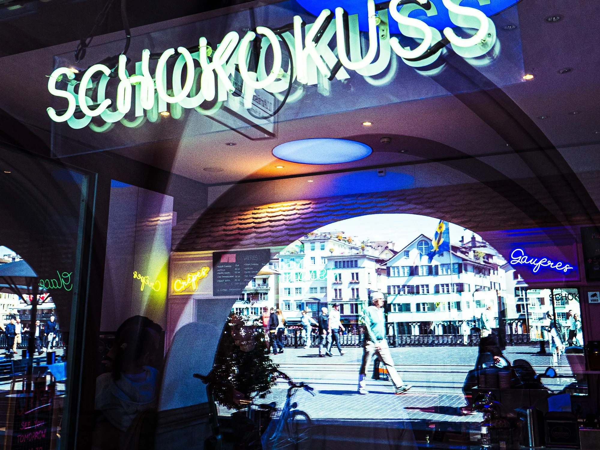 Bunte Neon-Schriftzüge im Fenster eines Cafés oder Restaurants mit Blick auf eine belebte Straßenszene im Hintergrund.
