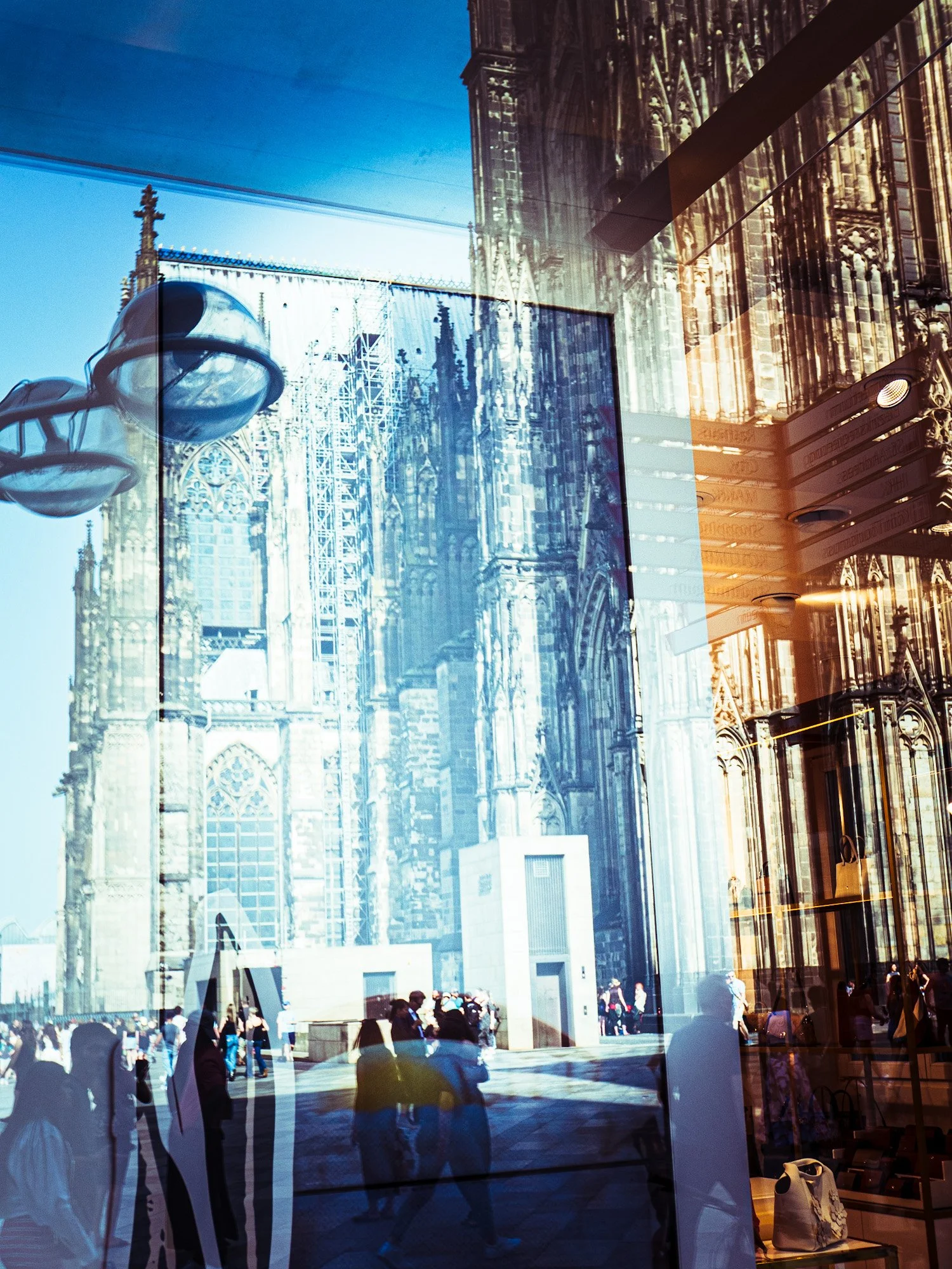 Innenansicht eines Gebäudes mit großen Glasfenstern, durch die eine Kathedrale mit gotischer Architektur und zahlreichen Menschen im Freien zu sehen ist.
