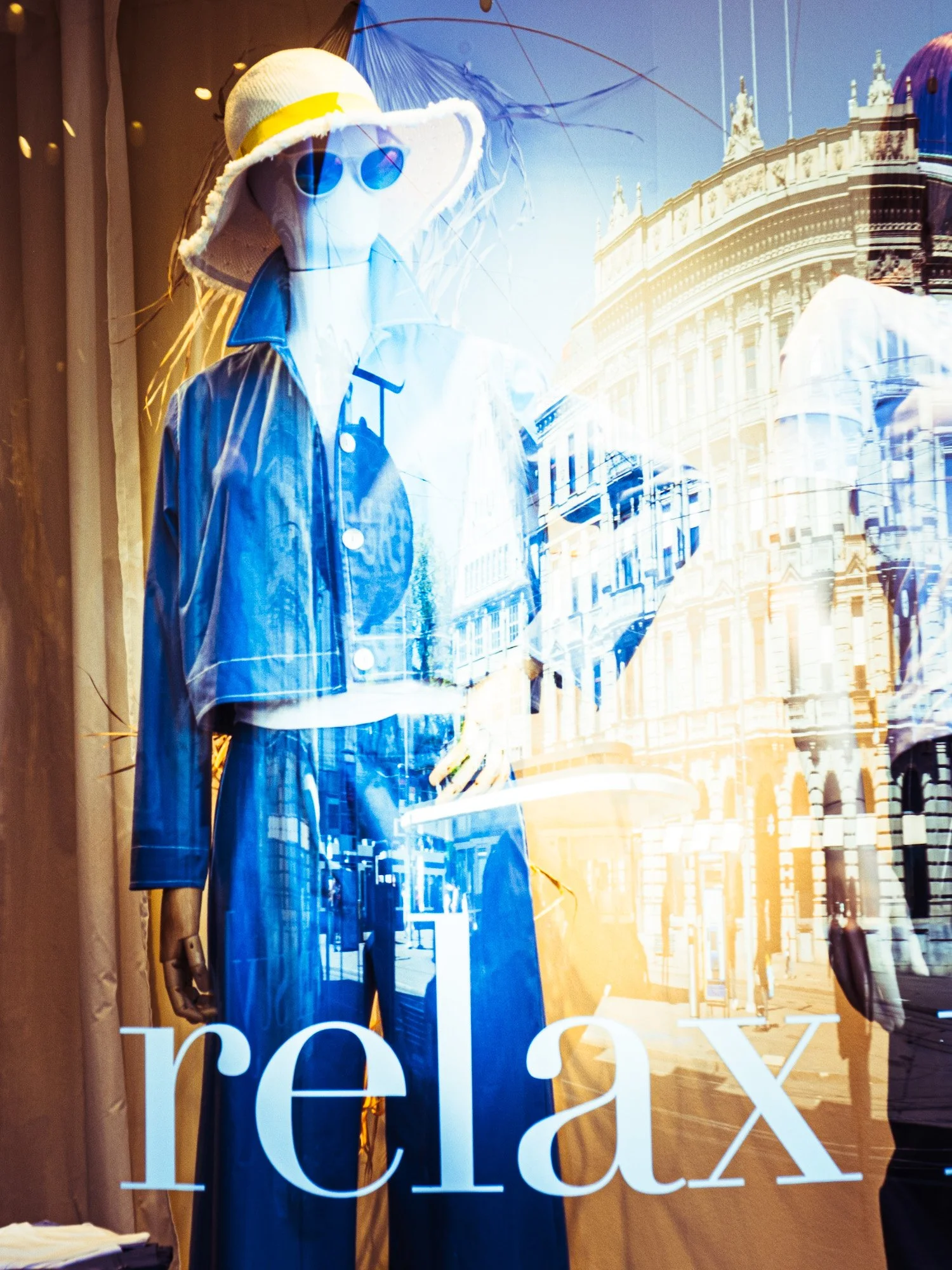 Schaufenster mit einer weiblichen Puppe in einem blauen Kleid und einem großen Sonnenhut, die durch eine Glasscheibe betrachtet wird, mit der Silhouette eines historischen Gebäudes im Hintergrund.