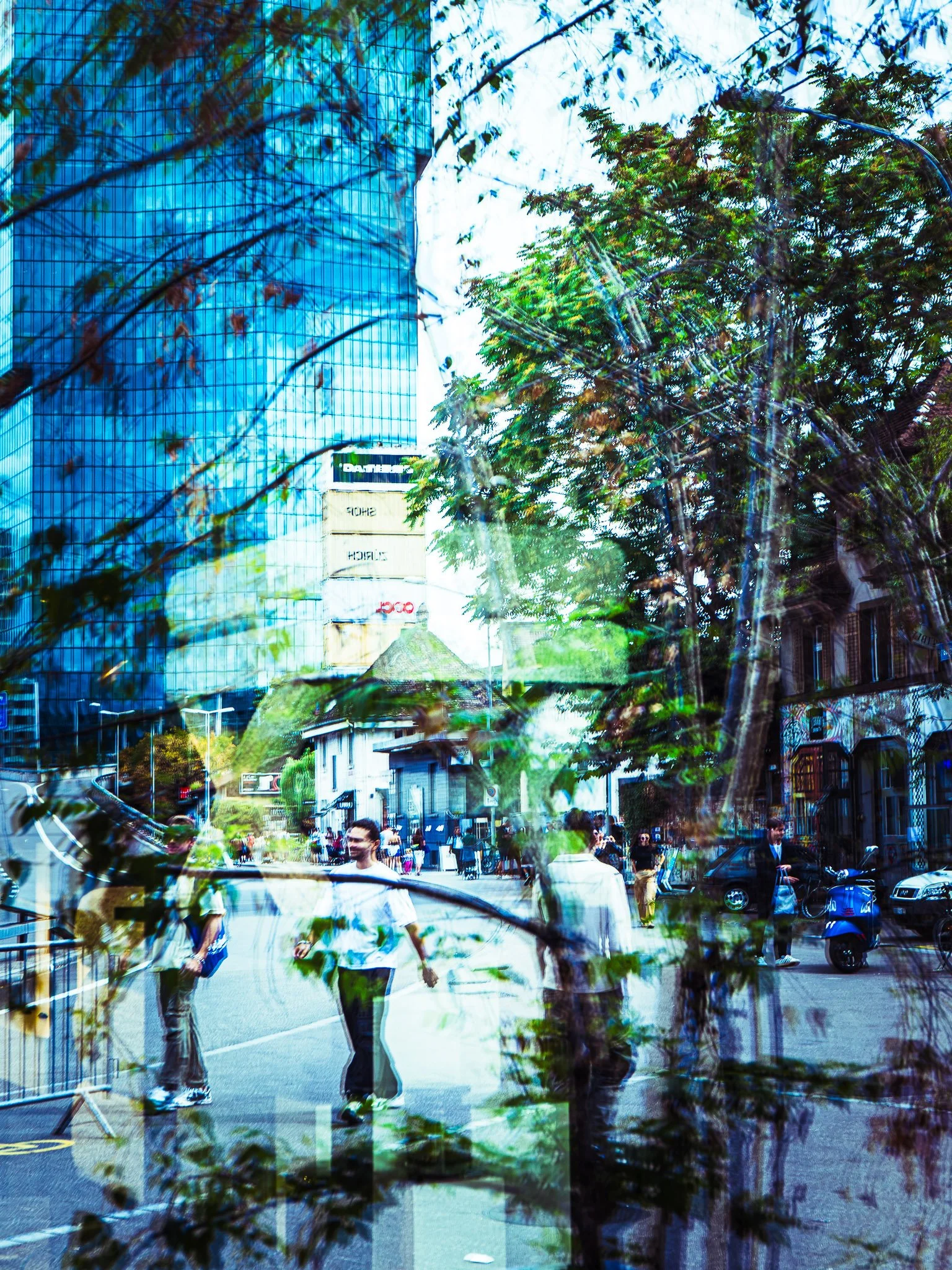 Window reflection in Zurich West: a blue glass skyscraper and stacked shipping-container tower merge with tree branches, while a smiling young man in a white T-shirt walks down a busy car-free street past a graffiti-covered building and a blue scoote