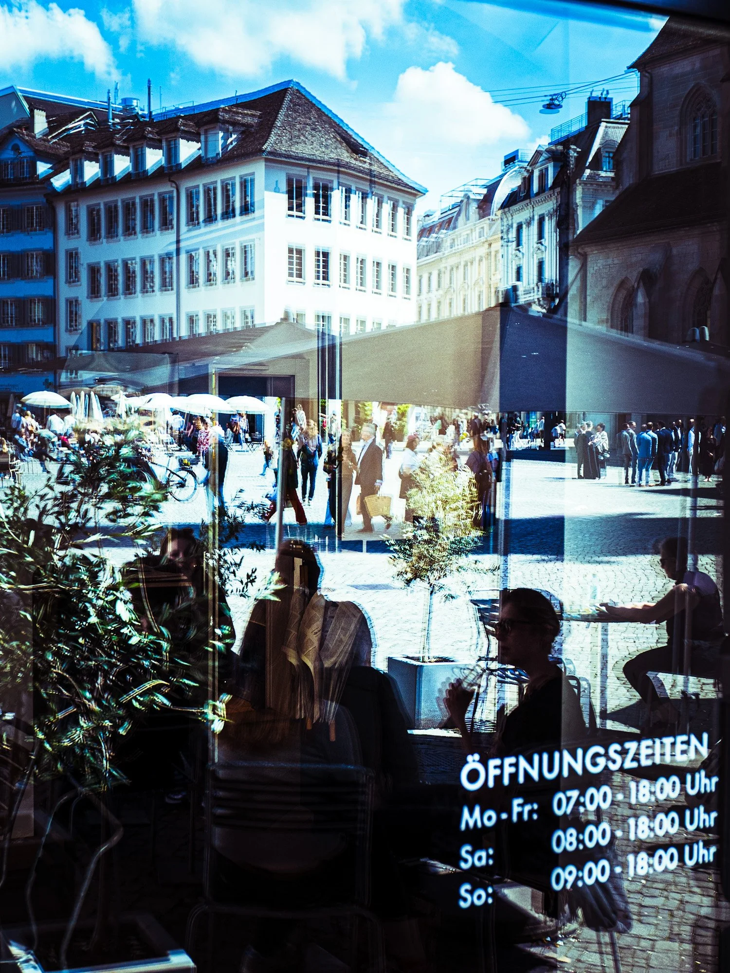 Ein Blick durch ein Fenster, das den Blick auf eine belebte Stadtstraße mit alten Gebäuden, Menschen, die spazieren gehen, und einem Markt mit Sonnenschirmen erlaubt. Im unteren Bereich sind Menschen im Inneren eines Cafés oder Restaurants zu sehen, 