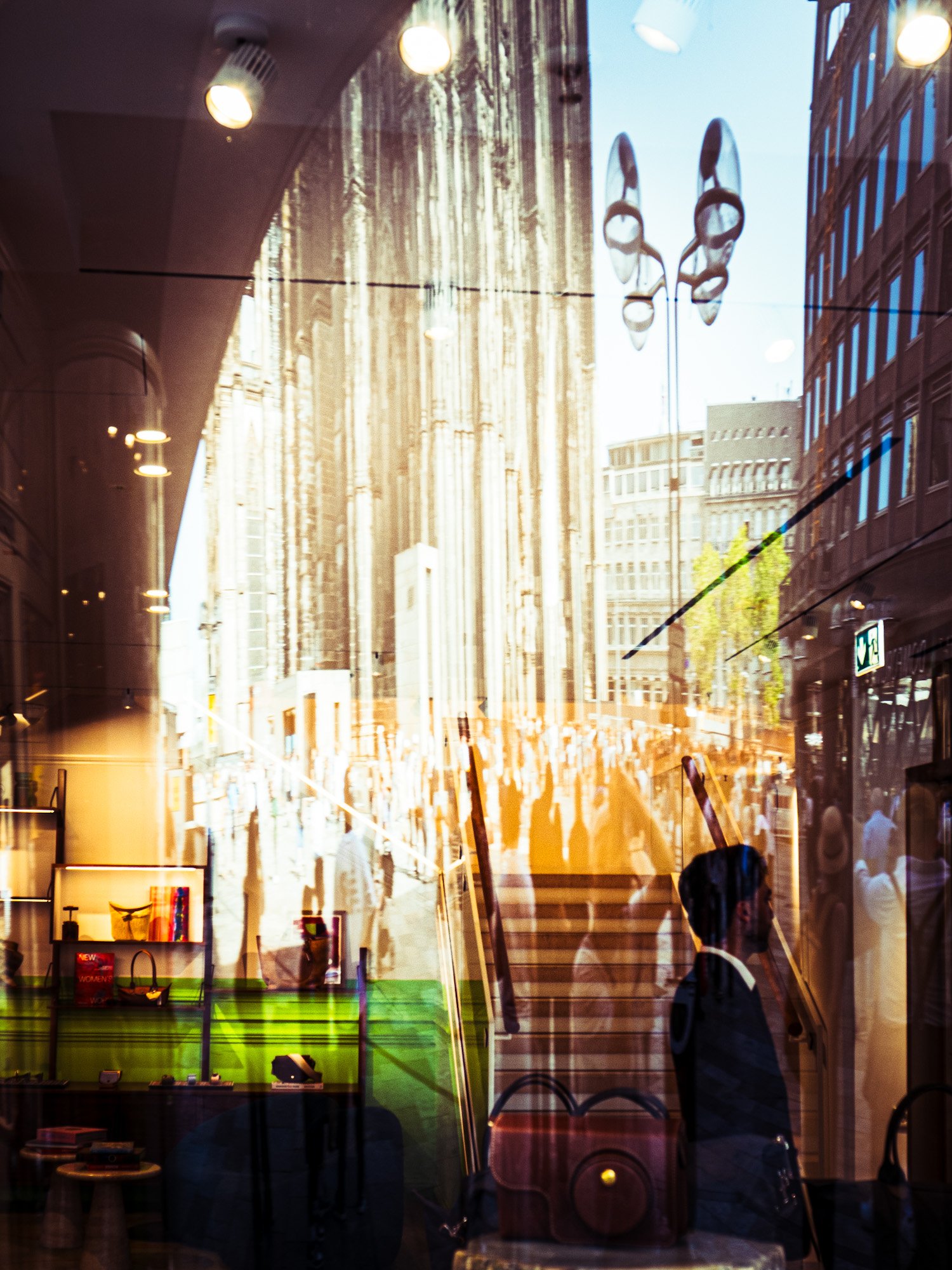 Innenaufnahme eines Geschäfts mit Schaufenster, reflektierende Glasfassade zeigt eine Stadt im Hintergrund. Im Vordergrund sind Regale und Ladentheke sichtbar, auf der einige Produkte stehen. Eine Person ist im Reflex im Glas zu sehen, sowie eine and