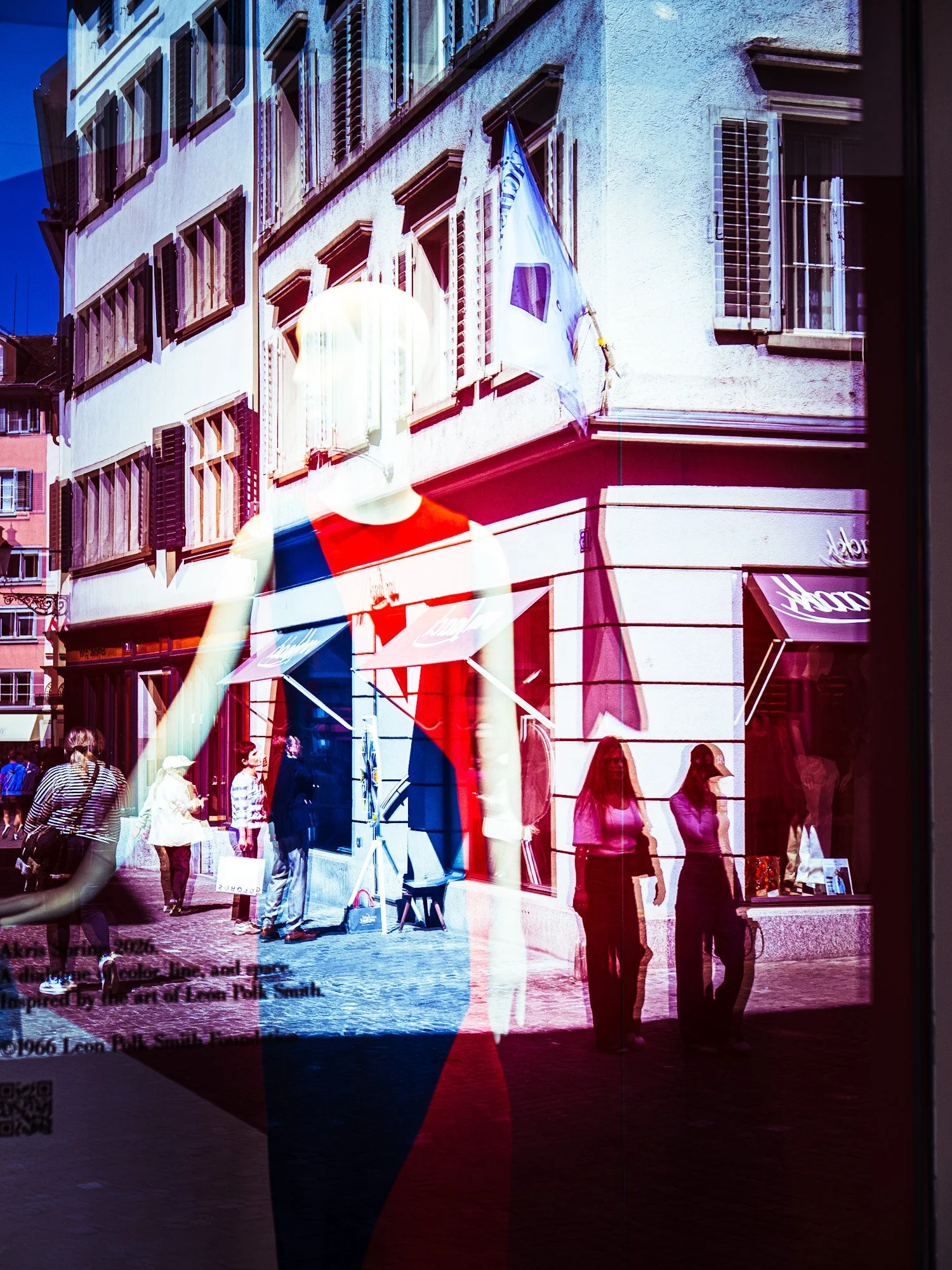 Reflexion eines Menschen in einem Schaufenster, draußen eine belebte Straße mit Menschen und Geschäften, alte Gebäude mit geschlossenen Fenstern, blauer Himmel.