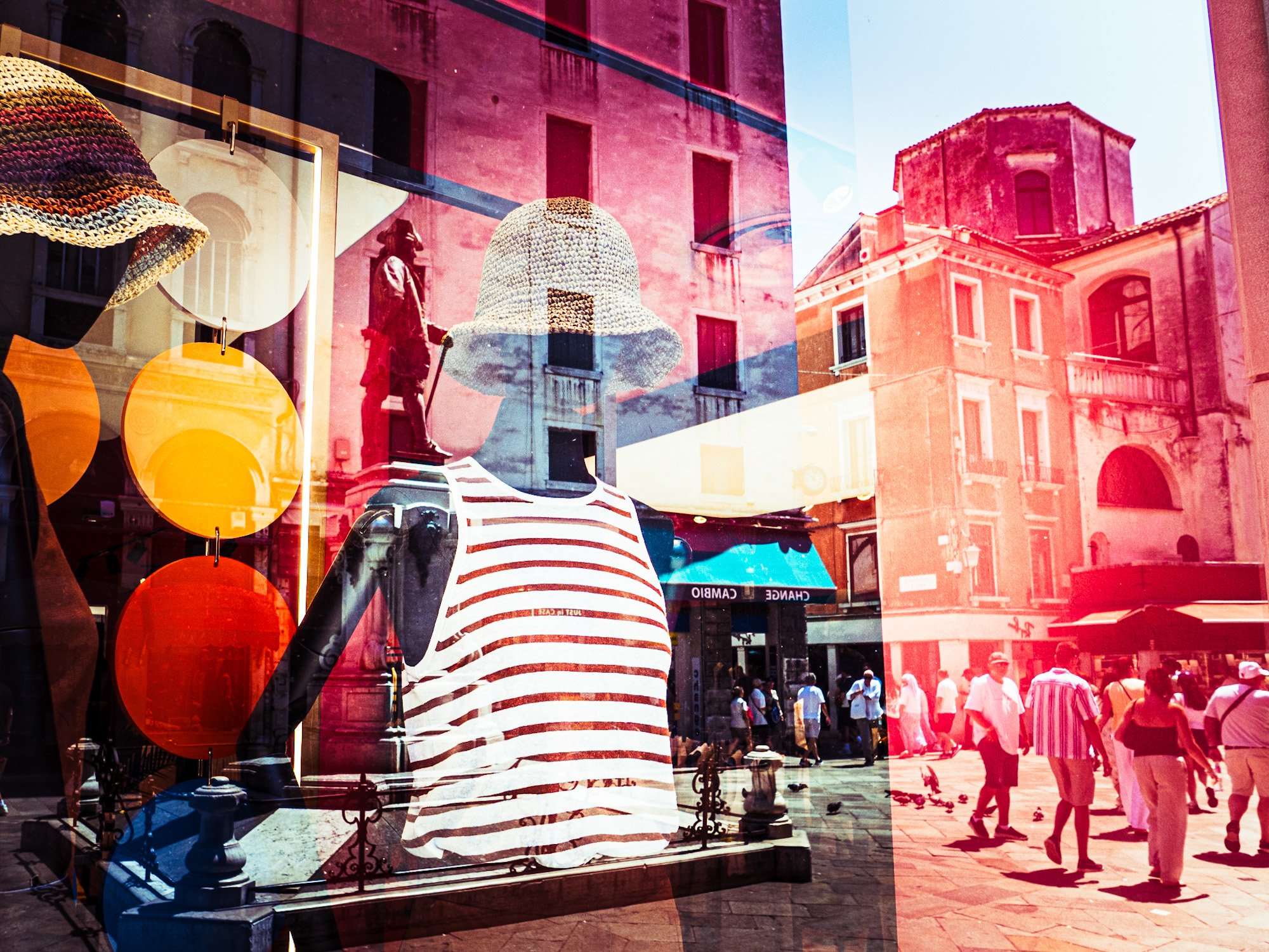Schaufenster mit bemalter Kleidung und Kopfbedeckung, reflektierende Gebäude und Menschen auf einem belebten Stadtplatz.