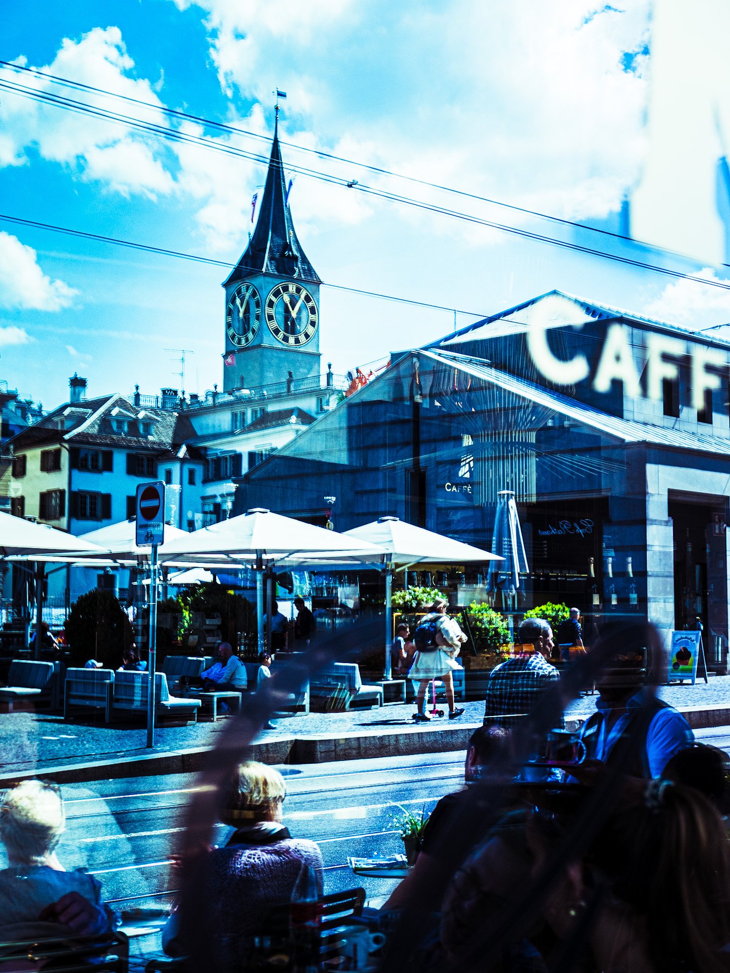 Blick auf ein Café im Freien mit Tischen und Stühlen, im Hintergrund eine alte Kirche mit einem Uhrturm und einer blauen Himmel mit Wolken.