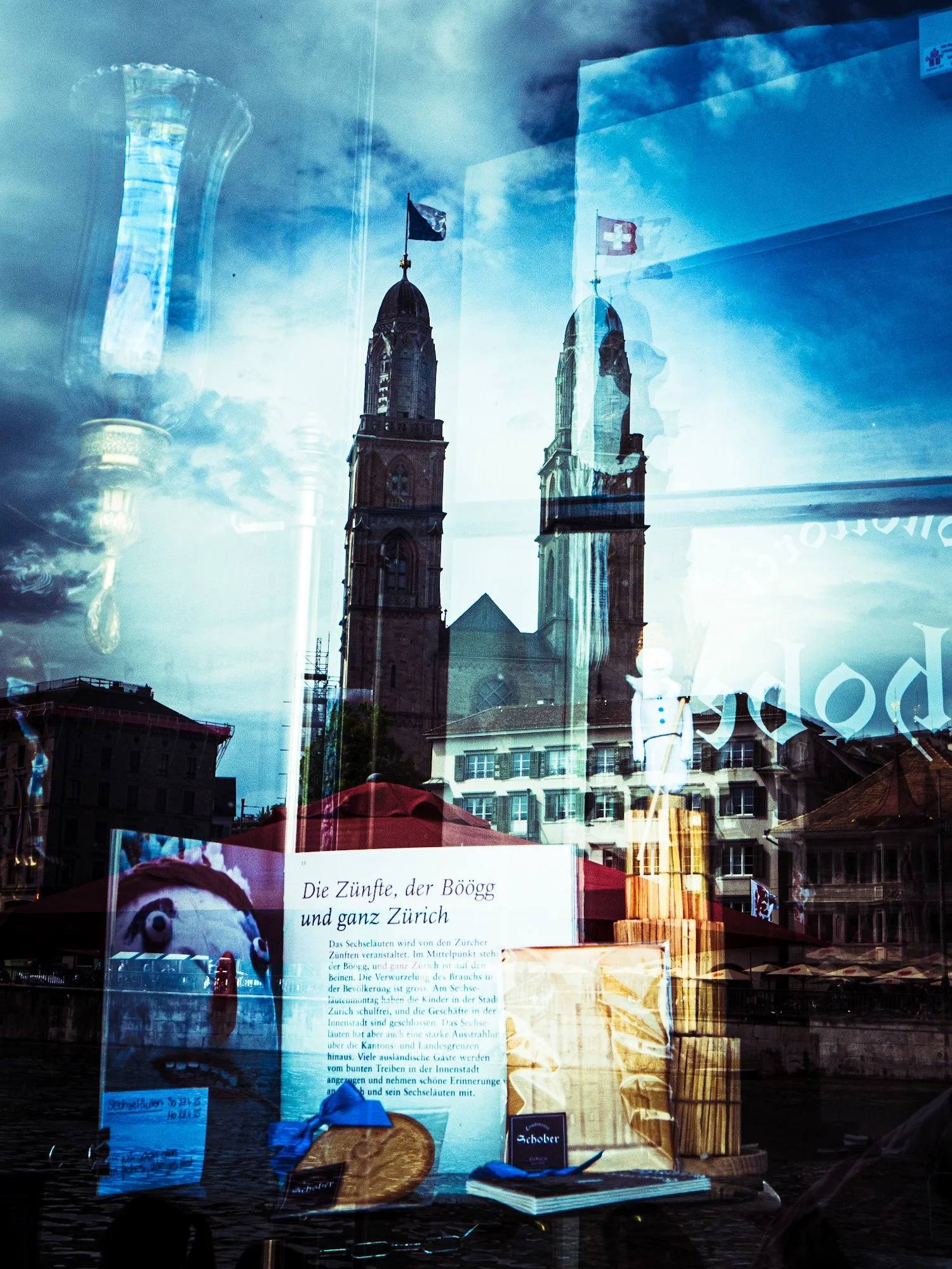 Reflexion eines historischen Kirchturms mit Flagge auf einer Glasscheibe, im Vordergrund sind Bücher, Poster und einen roten Schirm in einer Stadt, die Sommeratmosphäre vermittelt.