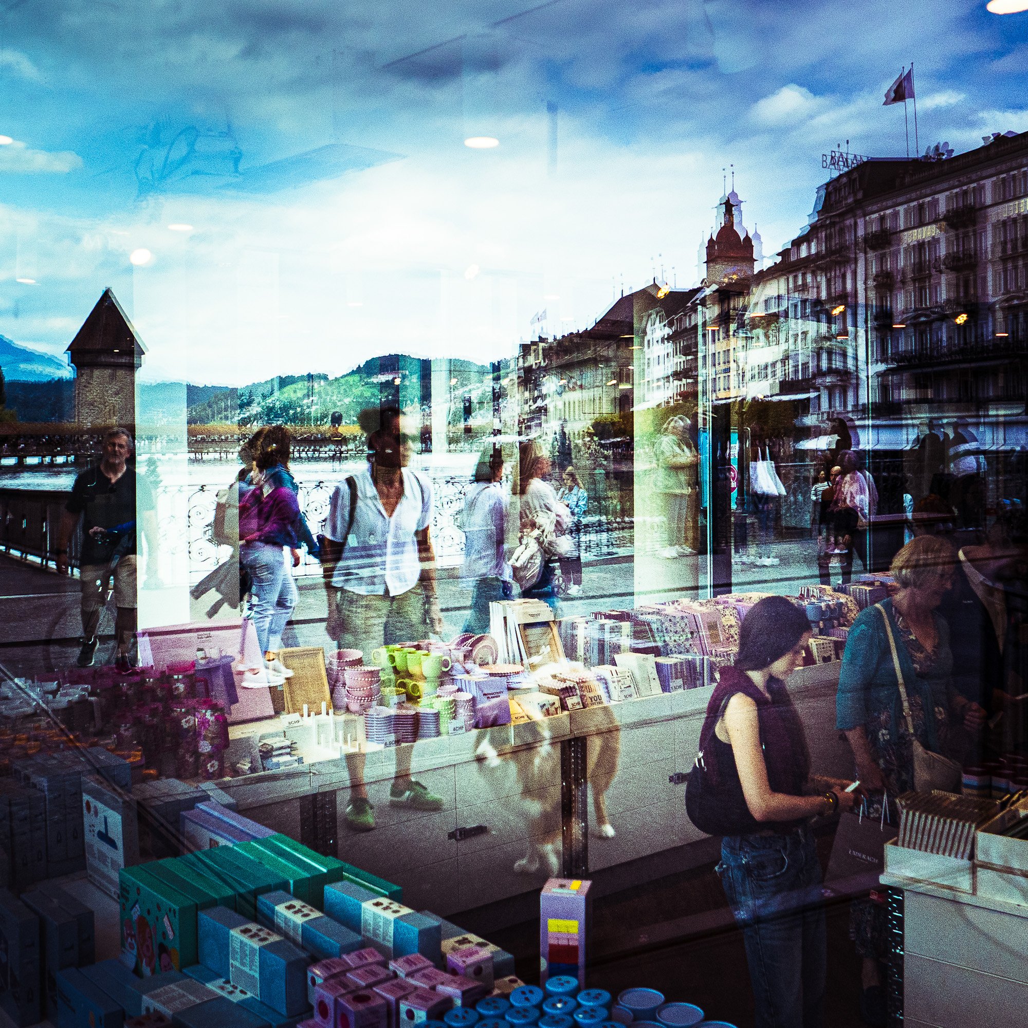 Blick durch ein Schaufenster auf einen Laden mit bunten Spielzeug- und Geschenkartikeln, Menschen gehen vorbei, im Hintergrund historische Gebäude mit Türmen und Flaggen, bewölkter Himmel.