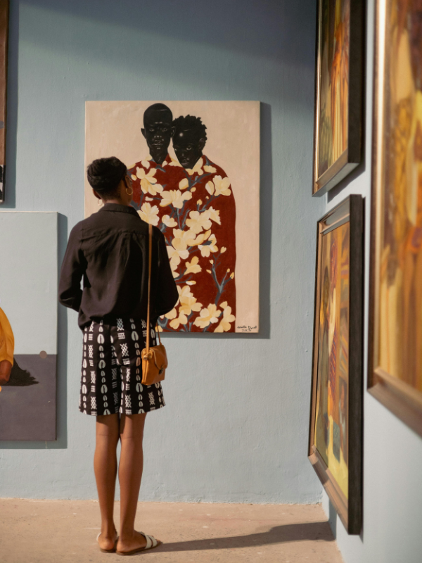 A woman standing in an art gallery observing a painting of two individuals with dark skin tones, with floral elements, hung on a light blue wall.