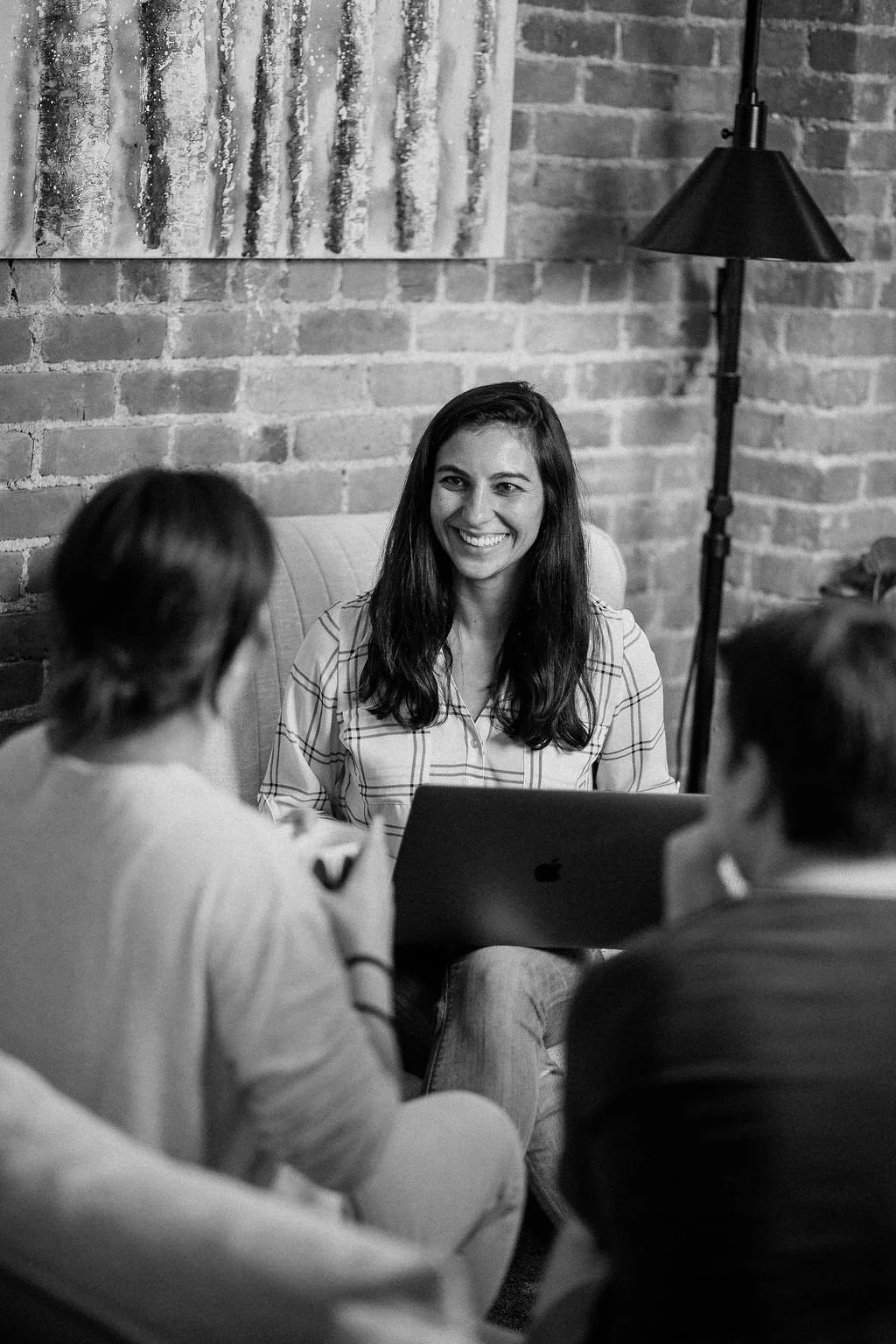 A group of four women sitting together in a modern coffee shop, engaged in a conversation. The woman in focus is smiling, wearing a striped shirt, with a laptop in front of her. The background features a brick wall and a decorative painting.