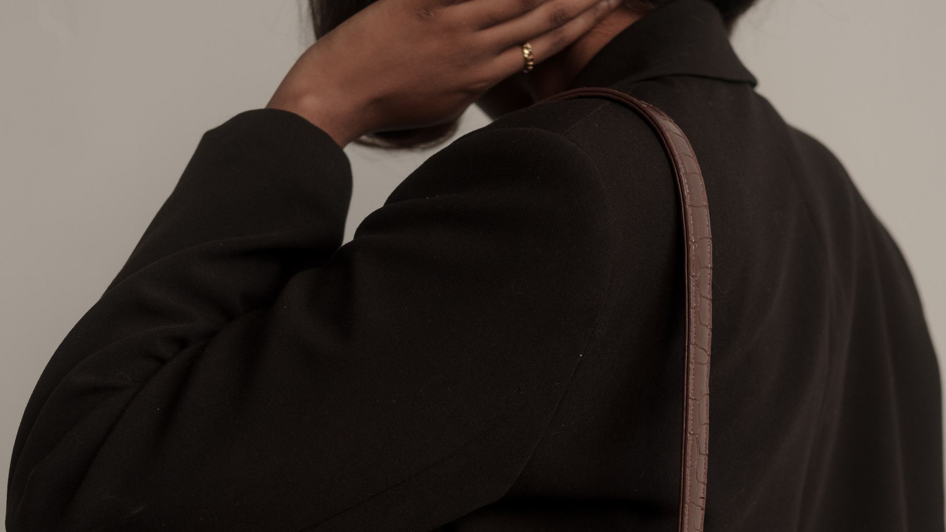 Close-up of a person wearing a black blazer, carrying a brown shoulder bag, and gently touching their face with one hand. The background is plain and neutral.