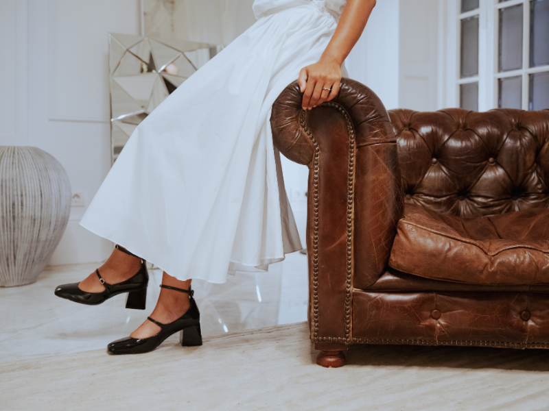 Person in a white dress and black high heels adjusting a brown leather armrest on a vintage sofa in a living room.