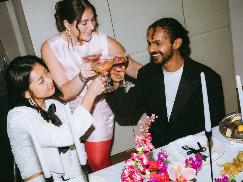 Three people celebrating with glasses of rosé wine at a dinner table decorated with pink and purple flowers, candles, and a fruit dish.