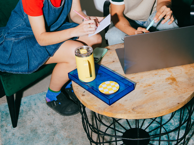 Two people sitting at a wooden coffee table working on a laptop and taking notes, with a yellow thermos, a striped coaster, and a phone on the table.