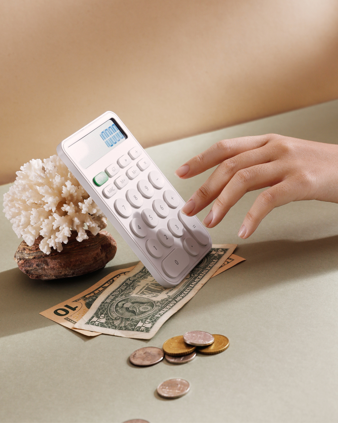 A hand pressing buttons on a white calculator placed on a dollar bill and coins, with a piece of coral on a rock as decoration, all on a green surface.