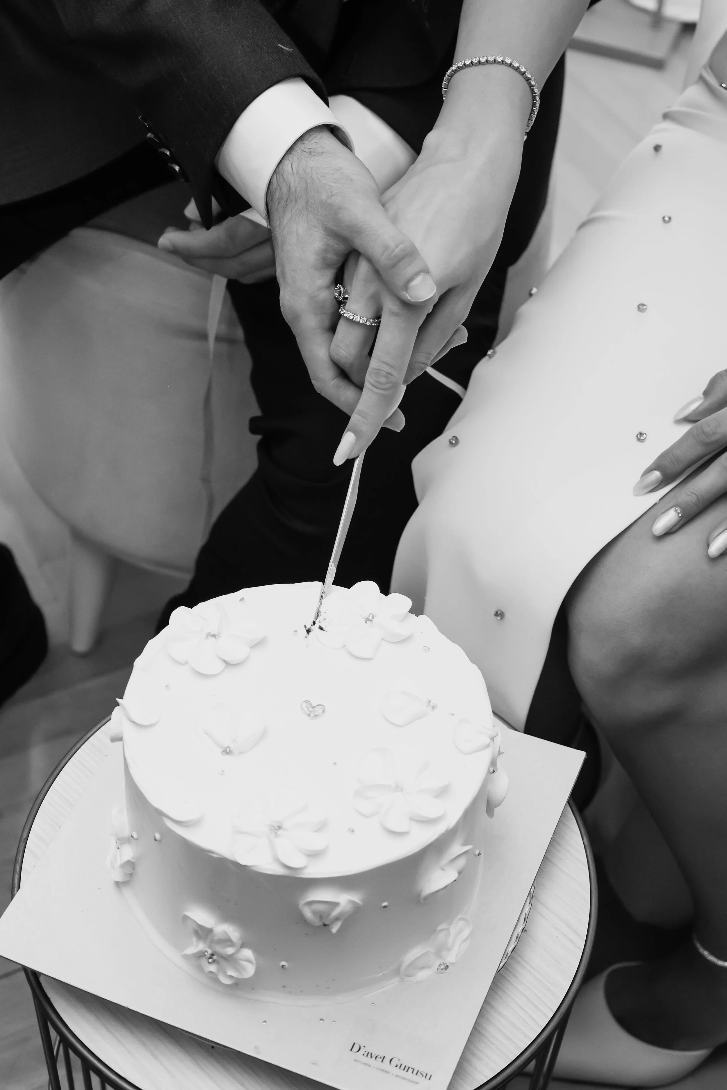 A couple's hands with wedding rings are intertwined over a white wedding cake decorated with small hearts and edible flowers. The groom is wearing a suit with a white shirt, and the bride is wearing a ring with a bracelet. The cake is on a plate with a card that has the text 'Davet Gunesi' on it.