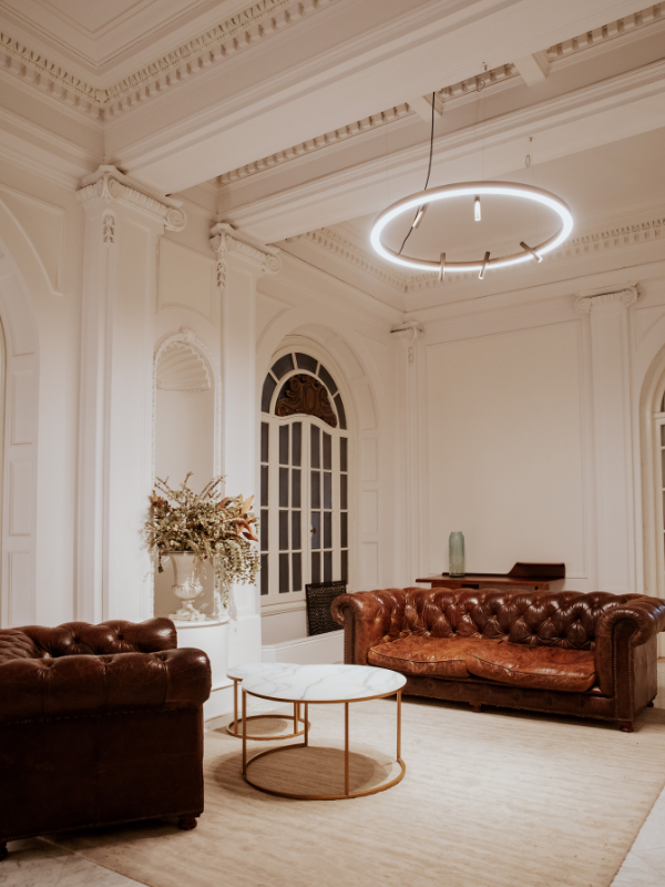 Living room with white walls, ornate crown molding, and large windows. Two brown leather sofas and a round coffee table with a white marble top. A tall, green decorative vase on a side table. Modern circular chandelier hanging from the ceiling.