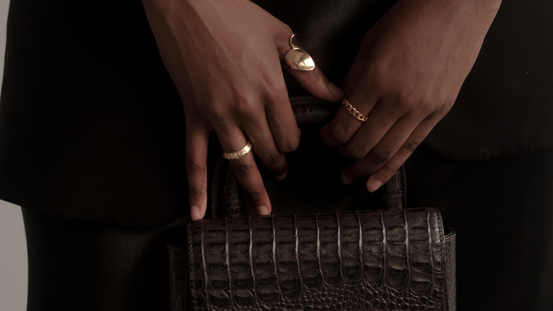 Person's hands resting on their waist, wearing multiple rings, holding a brown crocodile-patterned clutch bag, with black clothing in the background.