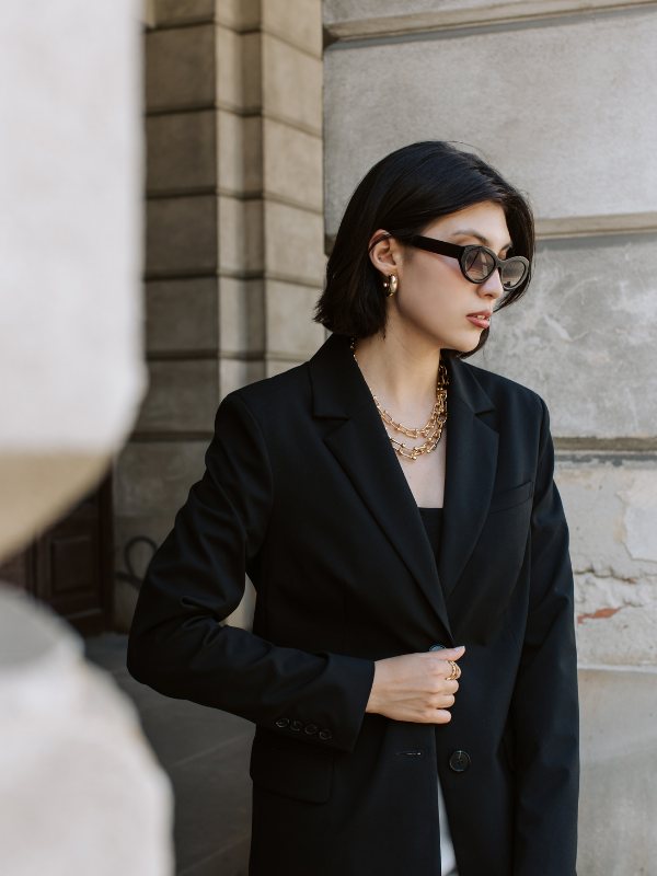 A woman with short black hair wearing dark sunglasses, gold jewelry, a black blazer, and a black top stands outside against a stone wall.