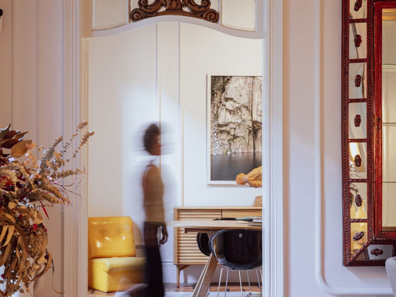 Blurred woman walking past a doorway in a decorated room with a yellow chair, framed artwork, and floral decor.