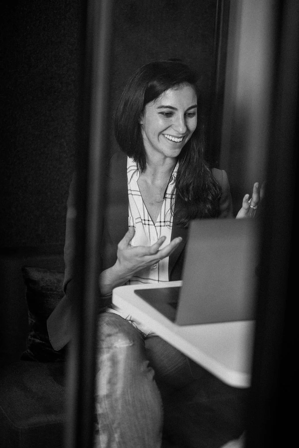 A woman with long dark hair, smiling, talking or demonstrating on a smartphone while sitting at a table with a laptop, through a partially visible door or curtain.