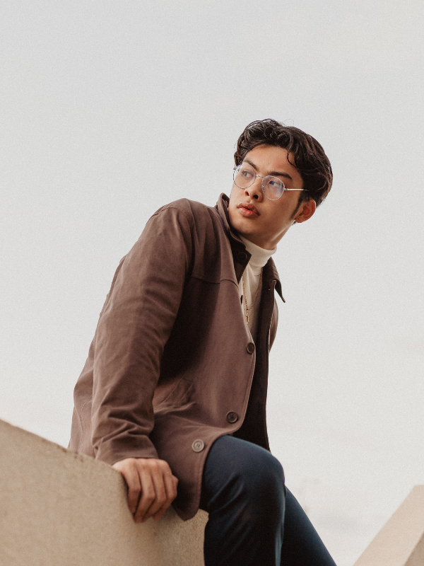 A young man with glasses wearing a brown jacket sitting on a concrete ledge against a gray sky