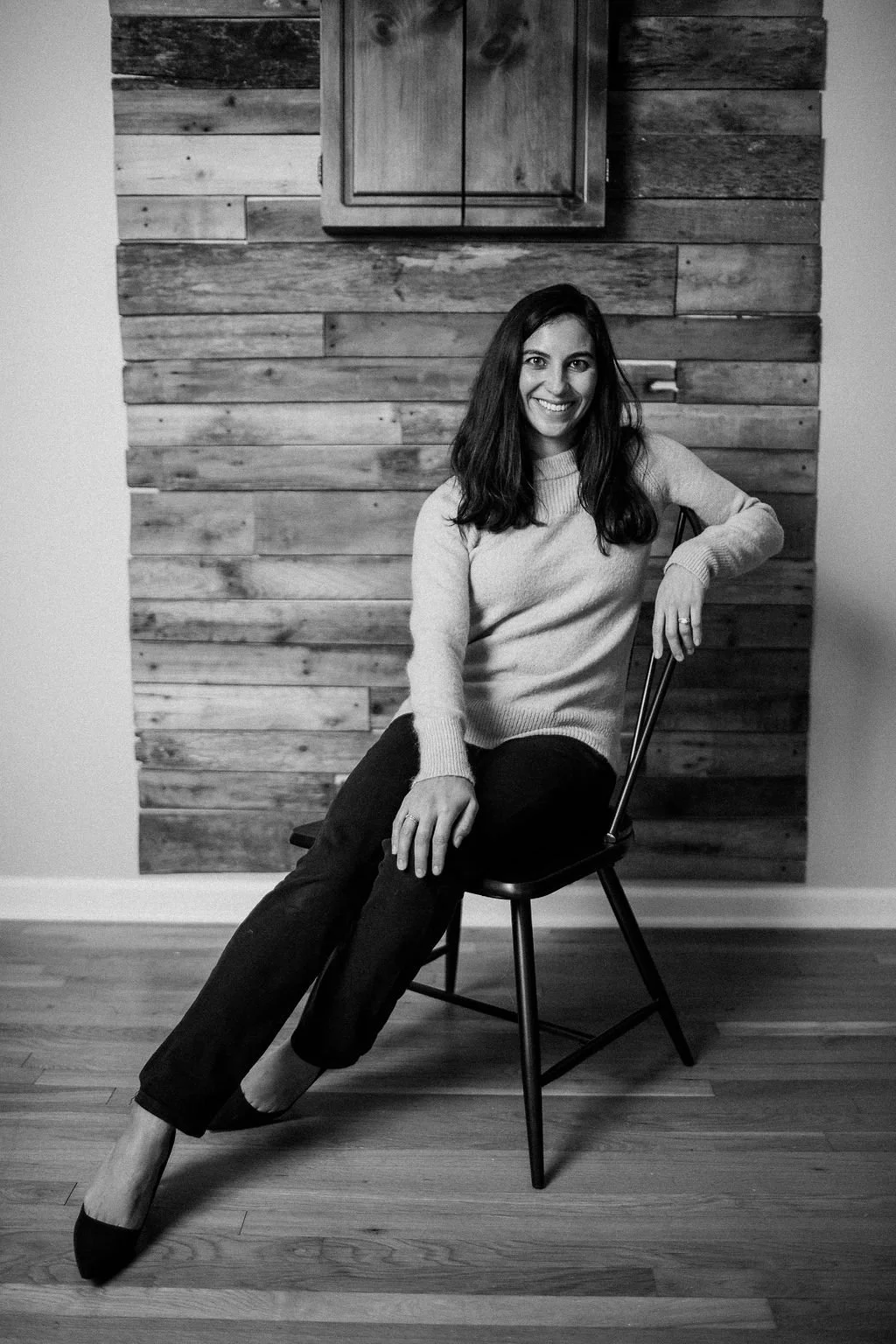 A woman with long dark hair wearing a sweater and pants, sitting on a chair and smiling in front of a wooden wall.