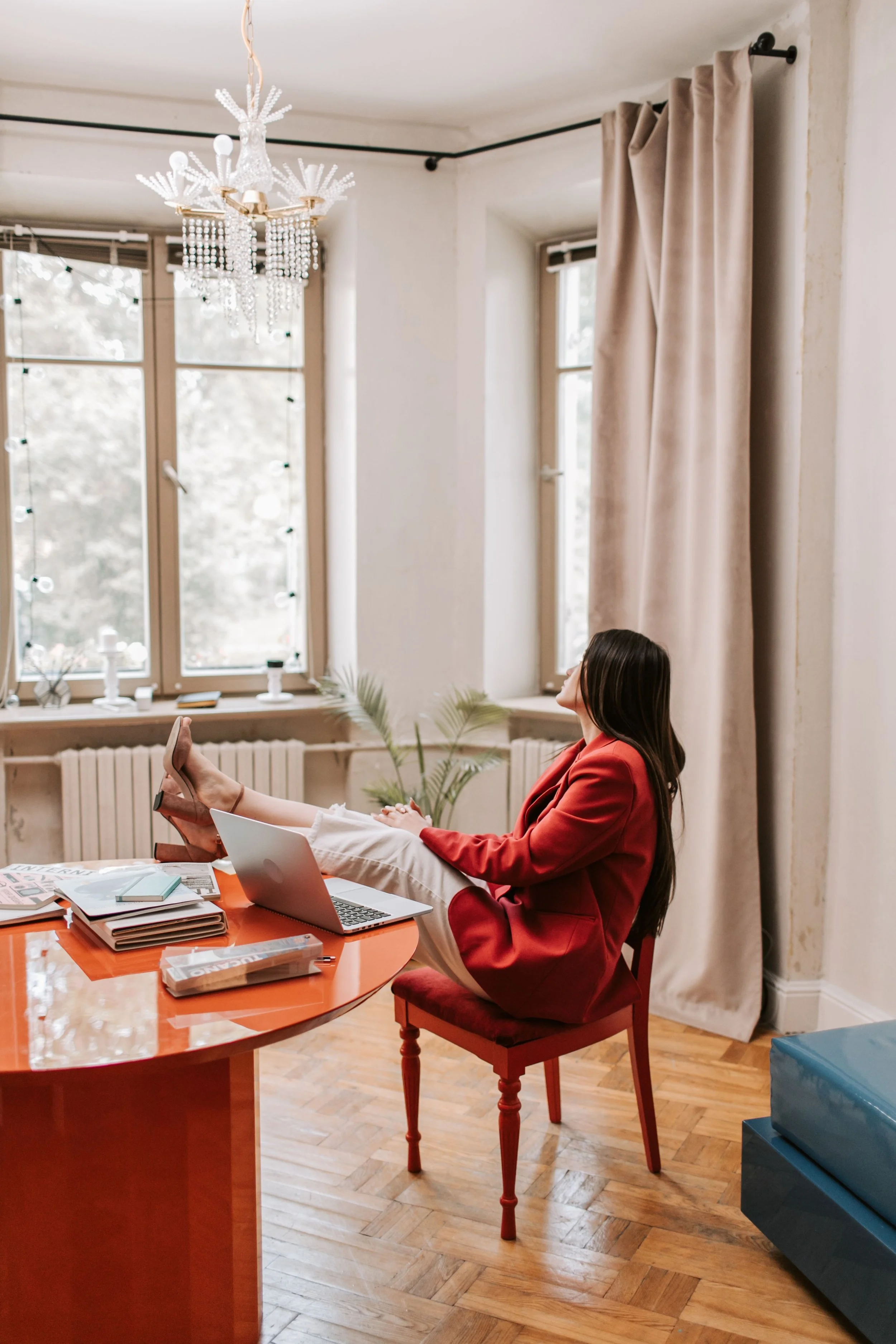 A woman in a red blazer and beige pants sitting in a room with hardwood floors, with her feet up on a table. She is wearing heels, and there are books, a laptop, and magazines on the table. The room has large windows with beige curtains, a chandelier, and some plants.